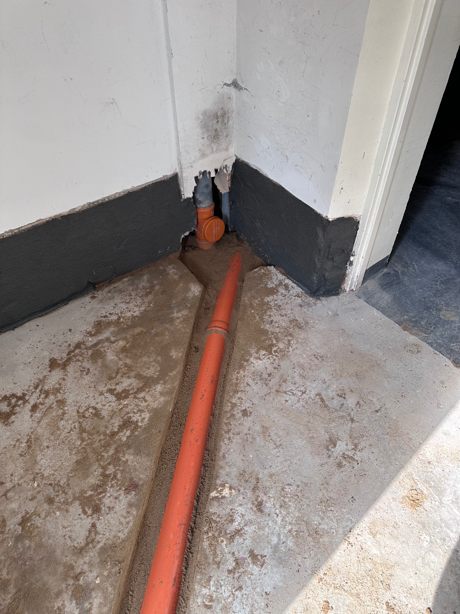 Pomarańczowa rura w wykopanym kanale w betonowej podłodze, przy ścianie z widoczną izolacją przeciwwilgociową oraz śladami wilgoci i pleśni.