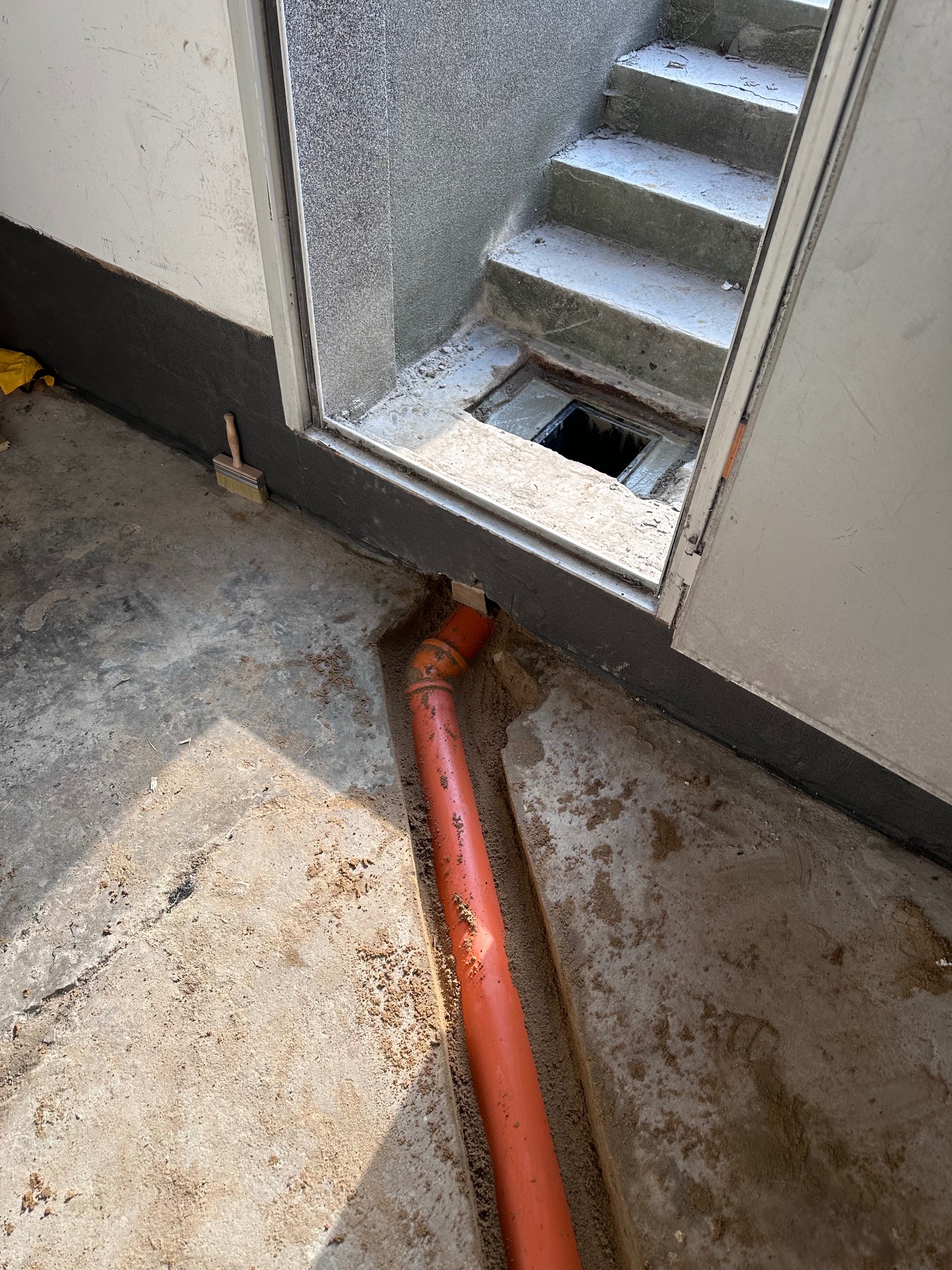 Pomarańczowa rura kanalizacyjna ułożona w wykopie w betonowej podłodze, widoczny fragment schodów i otwór kanalizacyjny, pędzel malarski oparty o ścianę.