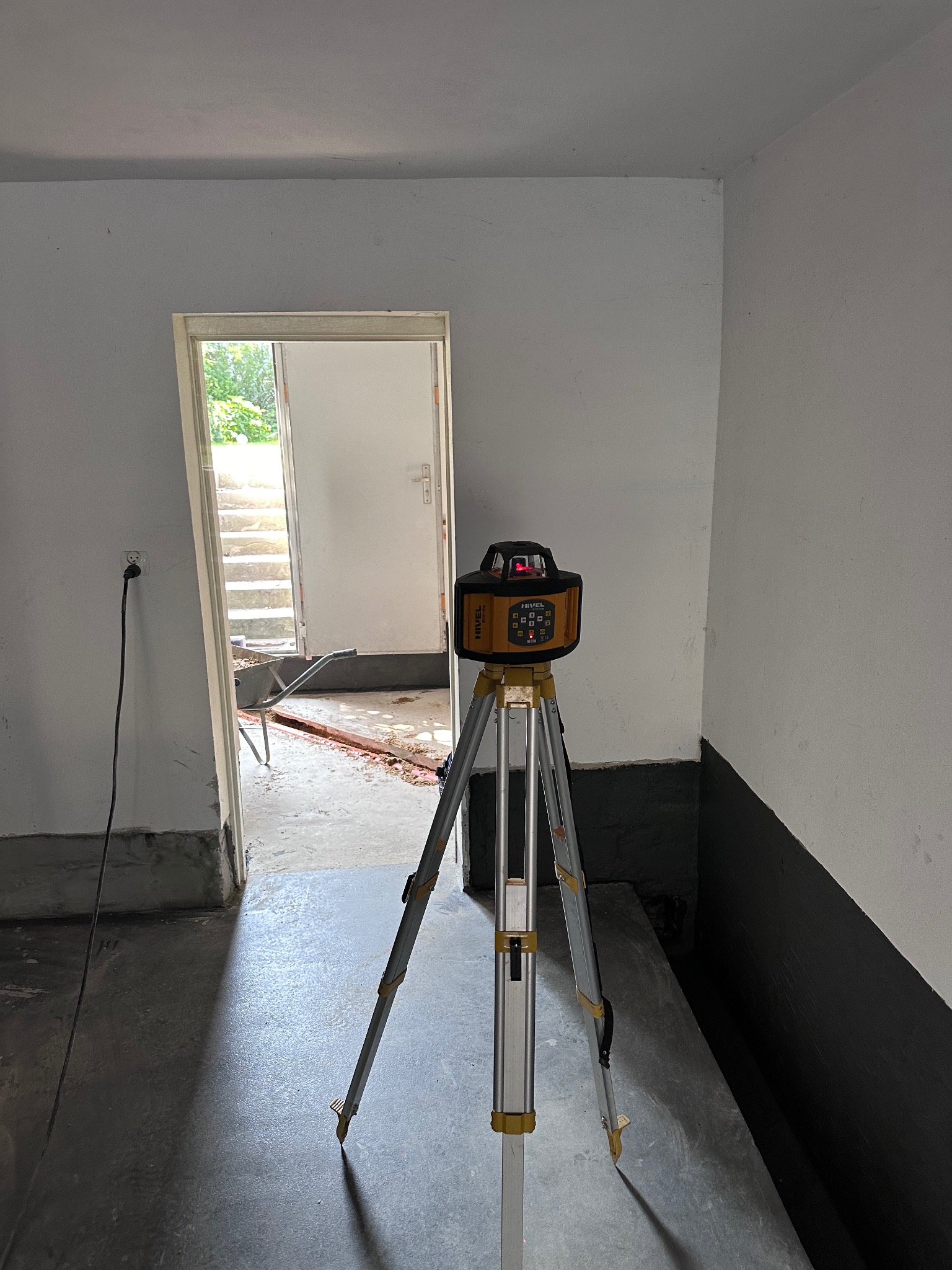 Poziomica laserowa na statywie ustawiona w surowym pomieszczeniu z widoczną bruzdą w podłodze i taczką w tle, sugerująca przygotowania do prac budowlanych.