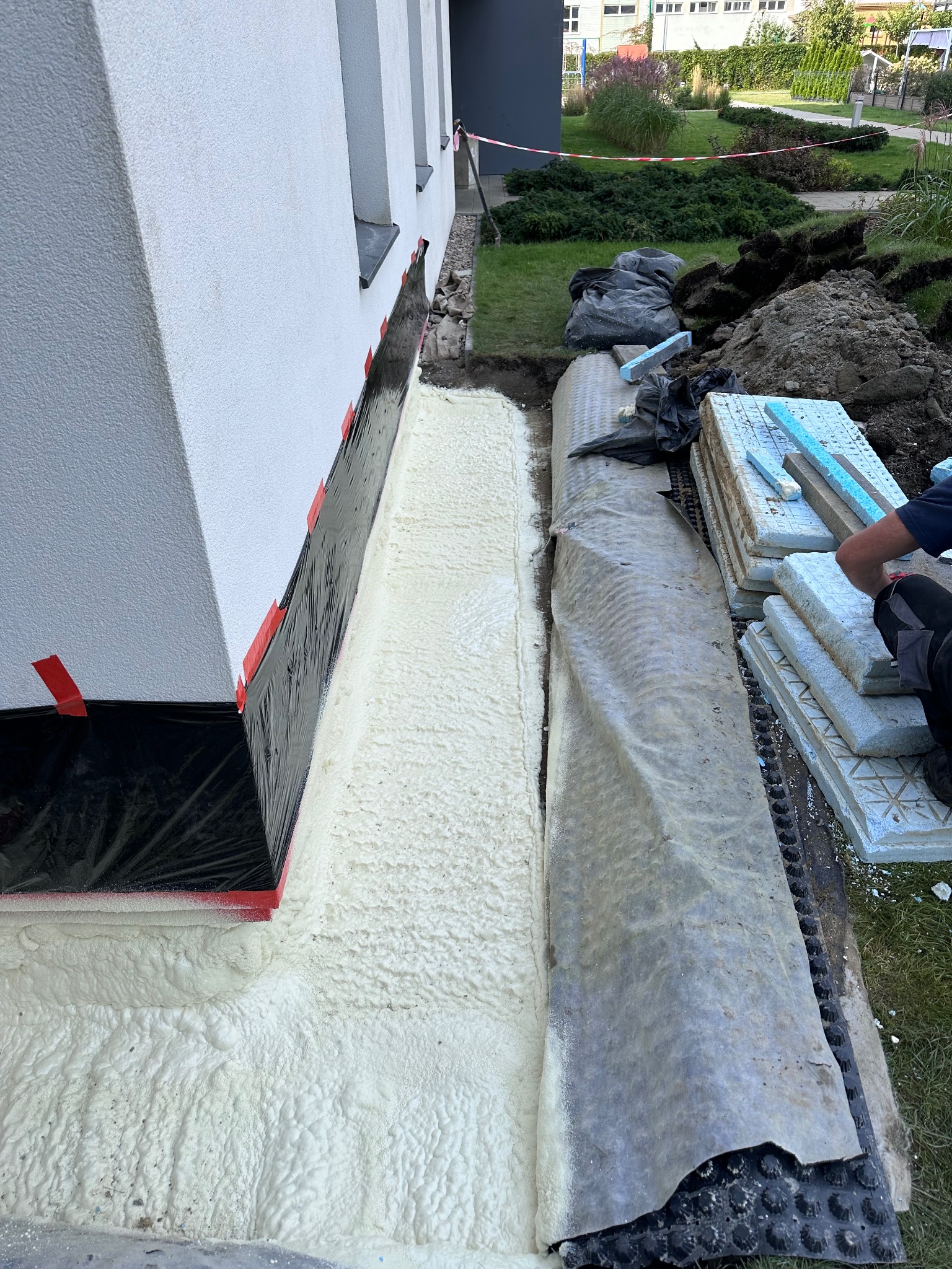 Aplikacja pianki izolacyjnej na fundamencie budynku, zabezpieczona folią, z widocznym drenażem i stosem płyt styropianowych obok wykopu.