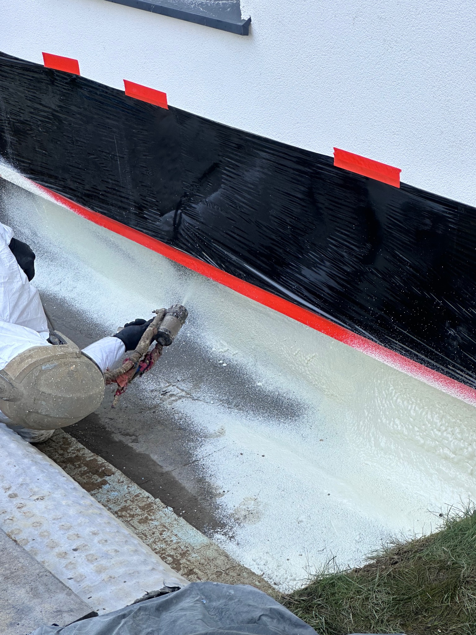 Natrysk pianki izolacyjnej na fundament zabezpieczony folią i taśmą, widoczny pracownik w kombinezonie ochronnym.