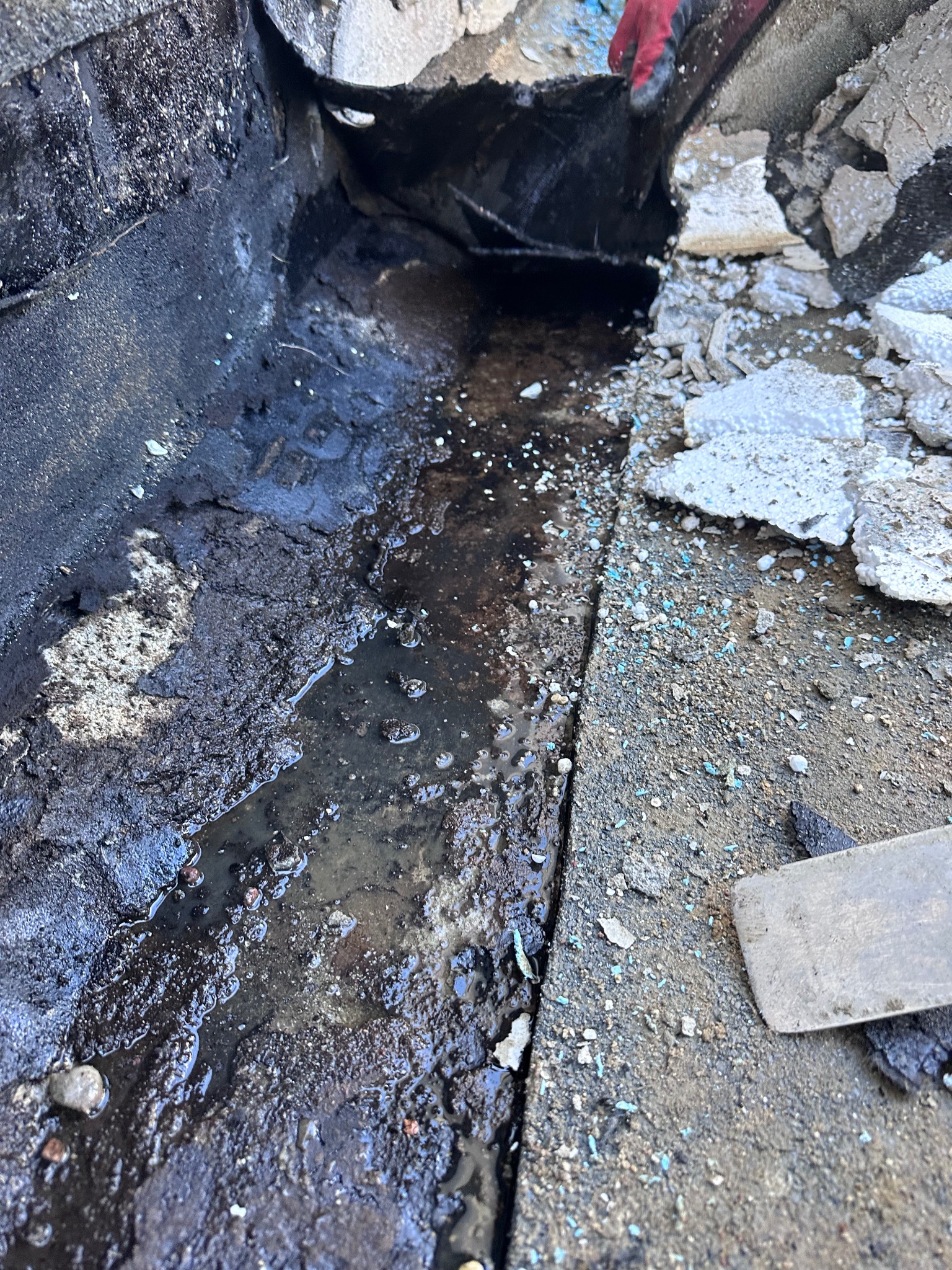 Zacieki wodne w wykopie fundamentowym, widoczne fragmenty izolacji i narzędzia, ziemia zanieczyszczona drobnymi elementami izolacyjnymi.