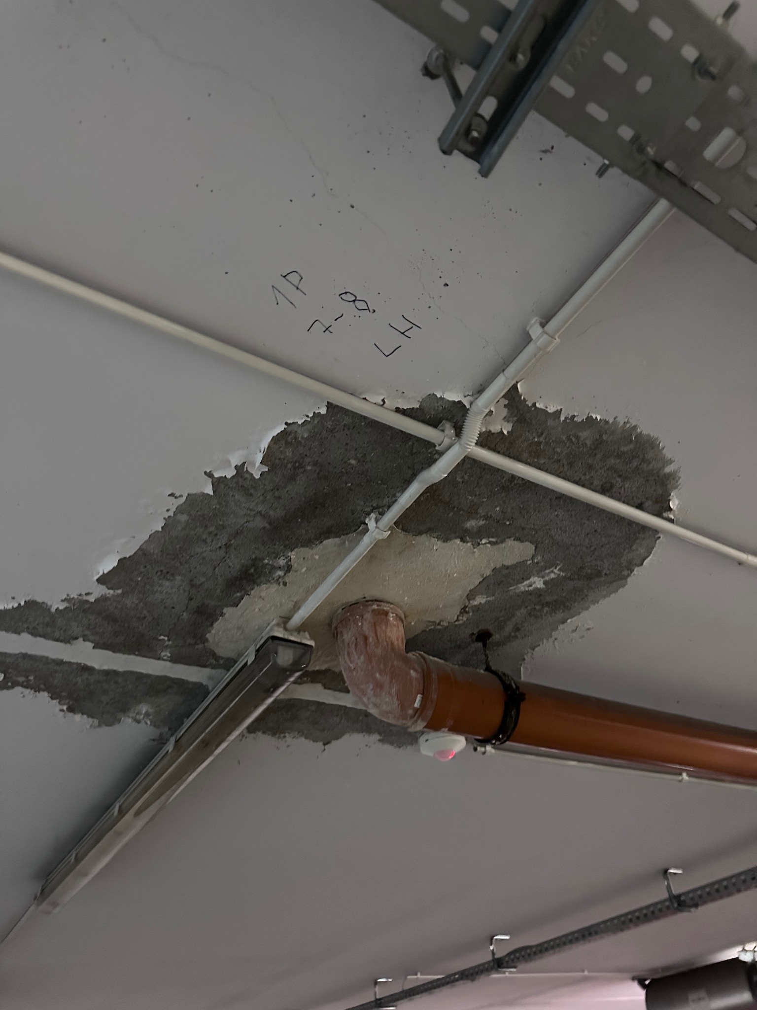 Uszkodzony sufit z widocznymi śladami wilgoci i napraw, rura kanalizacyjna z czujnikiem, instalacja elektryczna pod sufitem. Napis '1P 7-8 LH' na suficie.