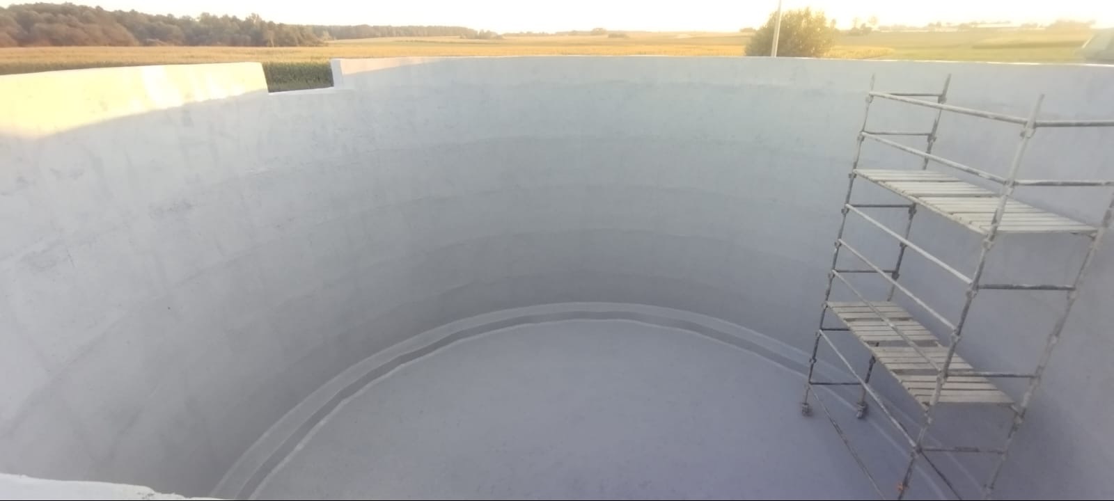 Wnętrze betonowego zbiornika, widoczne ślady po poziomych wzmocnieniach, z zewnątrz widok na pole, rusztowanie wewnątrz konstrukcji.