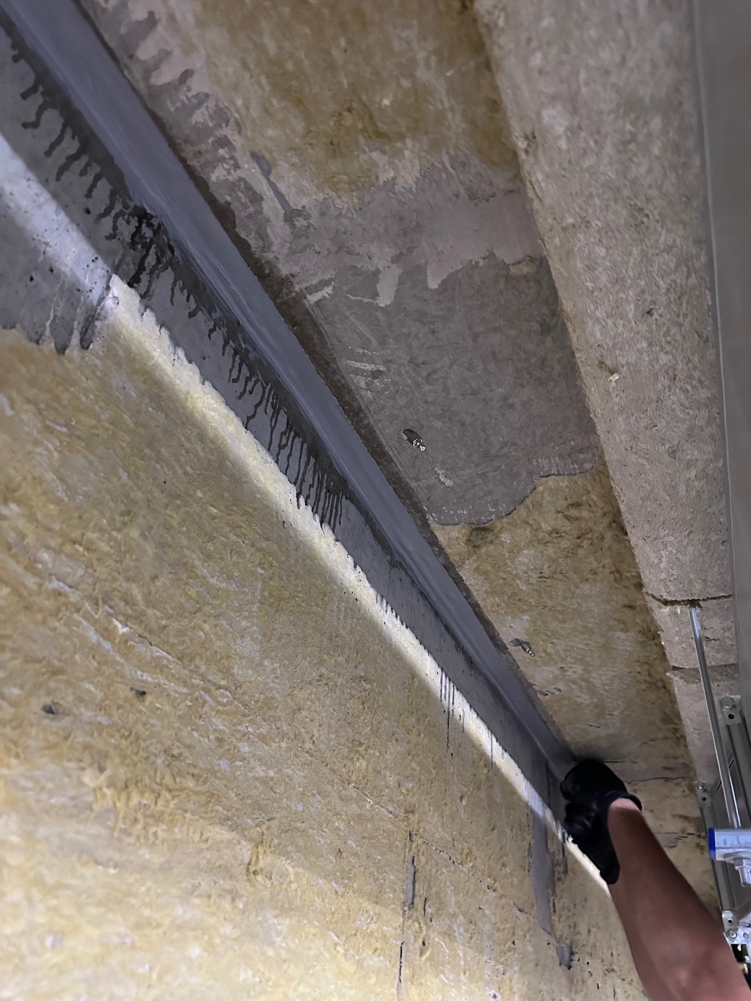 Aplikacja szarej masy uszczelniającej na styku betonowego fundamentu i izolacji z wełny mineralnej, widoczne ślady zacieków, praca w toku.