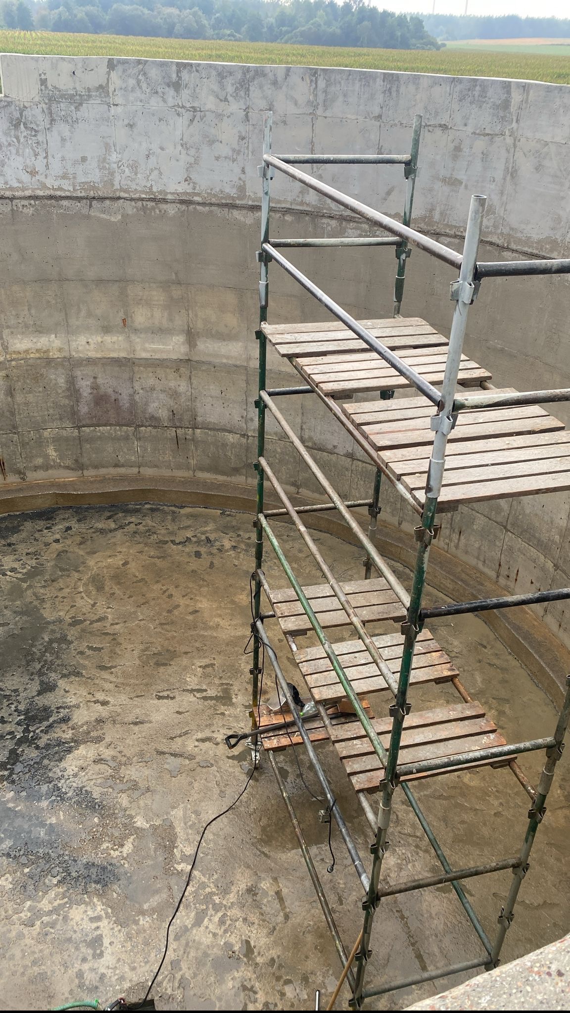 Wnętrze betonowego zbiornika z metalowym rusztowaniem, widoczne ślady wilgoci i zacieków na ścianach i podłożu, na zewnątrz zielony krajobraz.