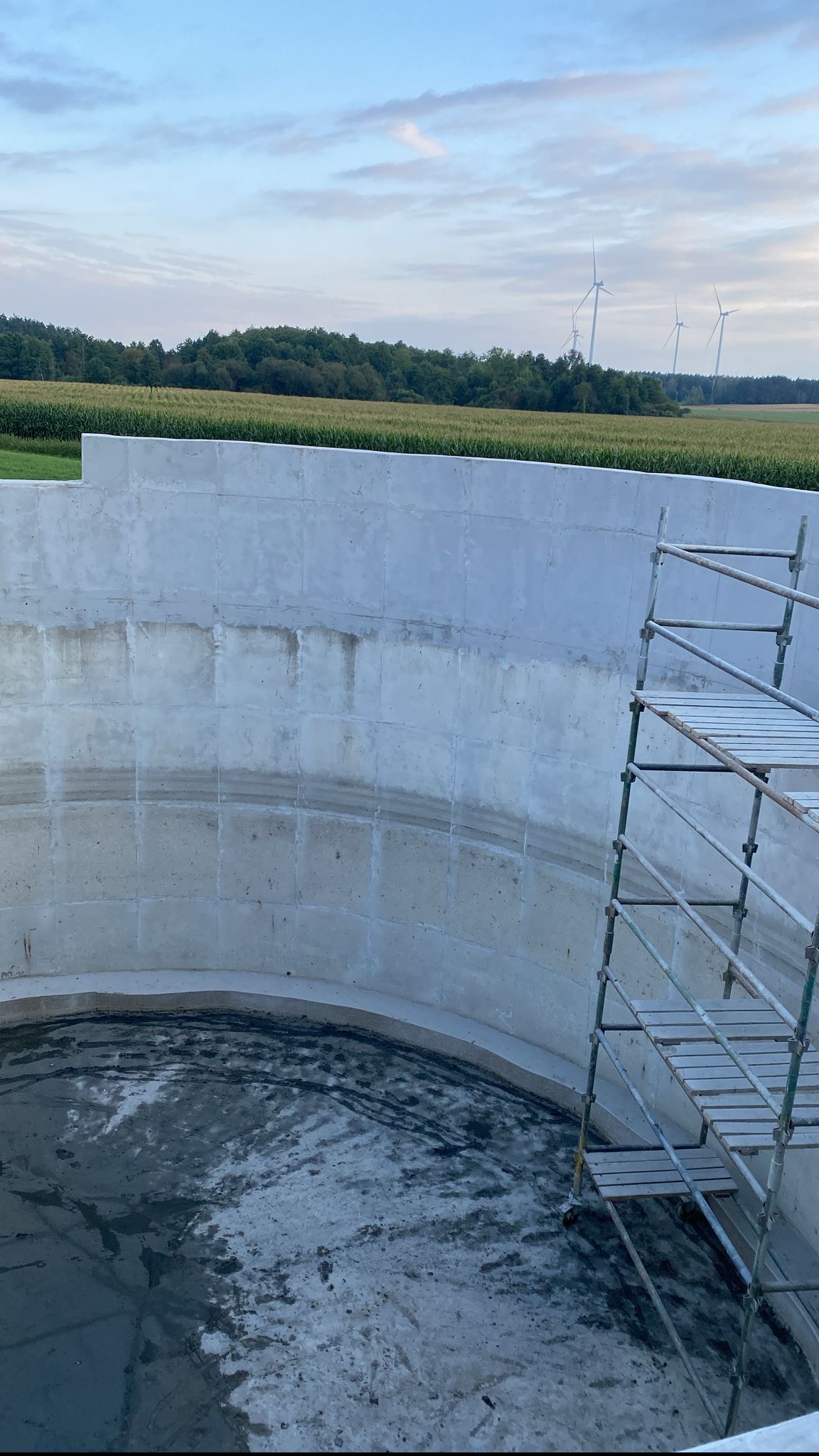 Betonowy okrągły zbiornik w trakcie budowy, widoczne ślady po wodzie, rusztowanie, w tle pole uprawne i turbiny wiatrowe.