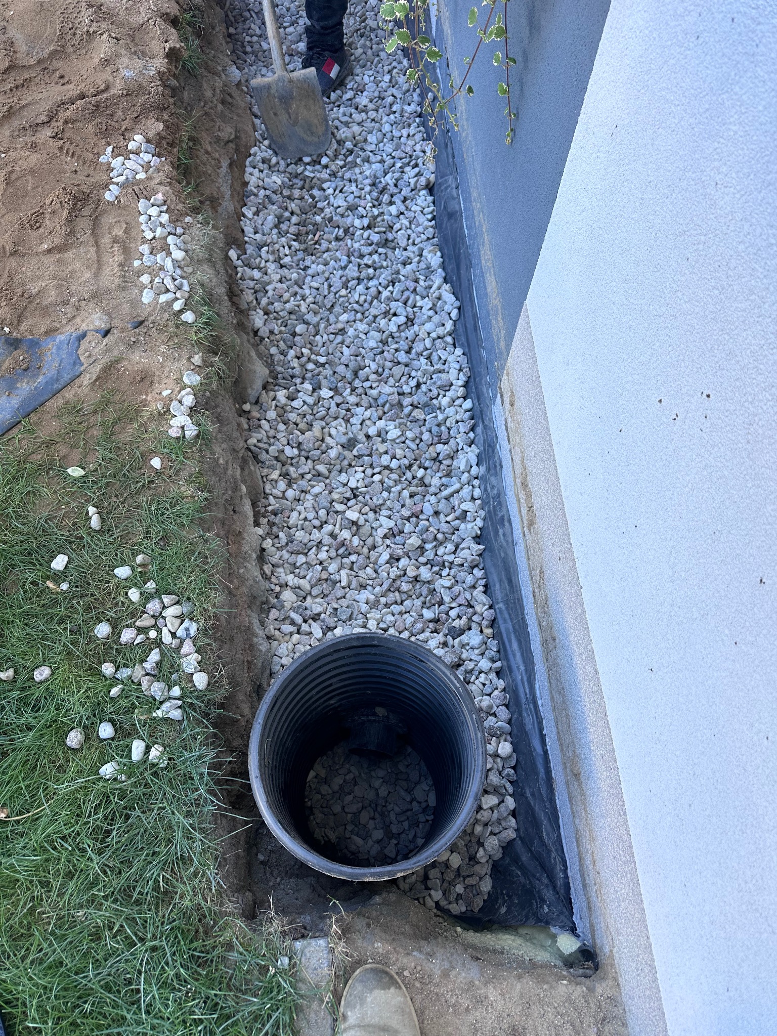 Wykop wzdłuż fundamentu budynku wypełniony drenażem z rurą odpływową, zabezpieczony czarną folią hydroizolacyjną.