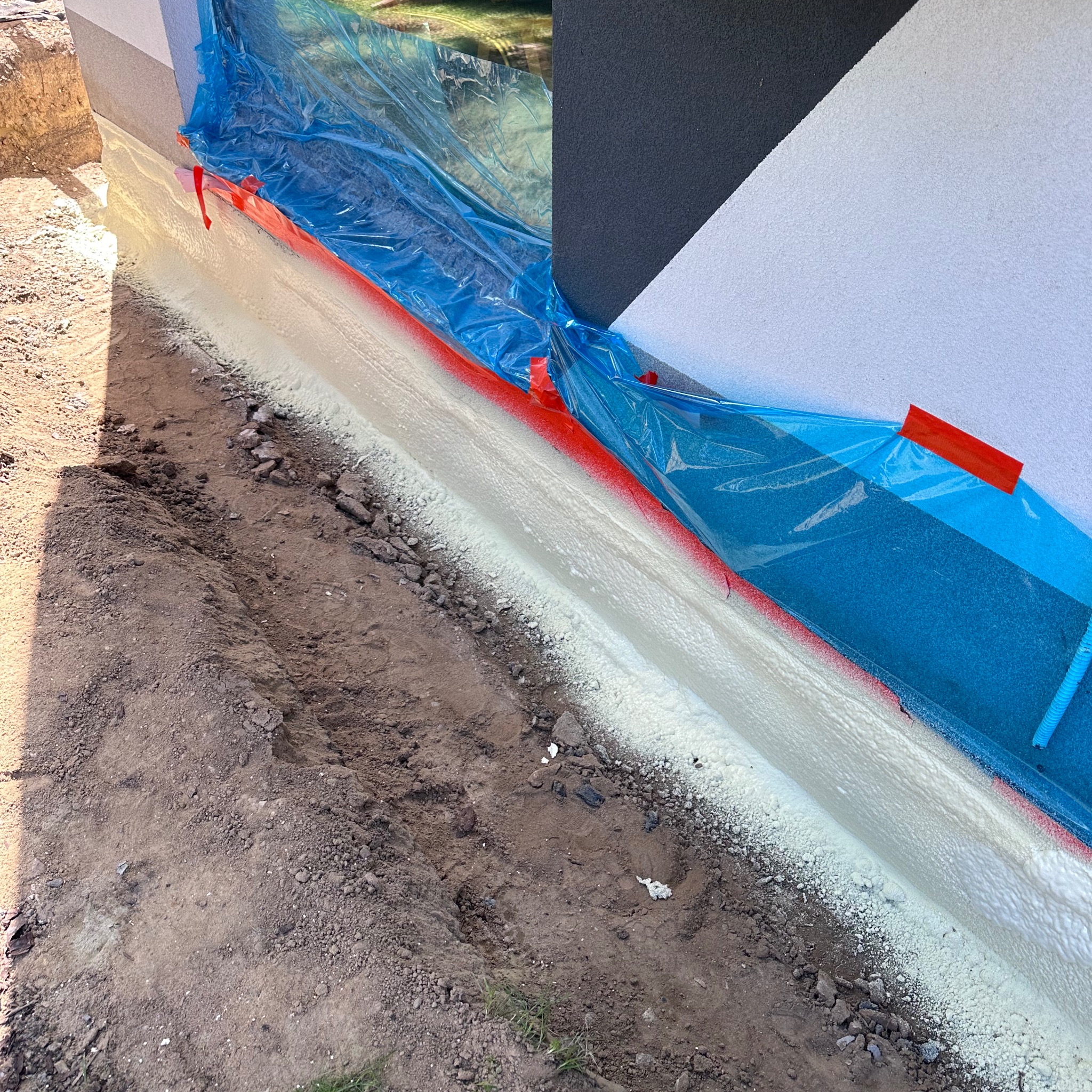 Świeżo nałożona, biała pianka izolacyjna na fundamencie budynku, zabezpieczona folią i czerwoną taśmą malarską, widoczna wykopana ziemia.