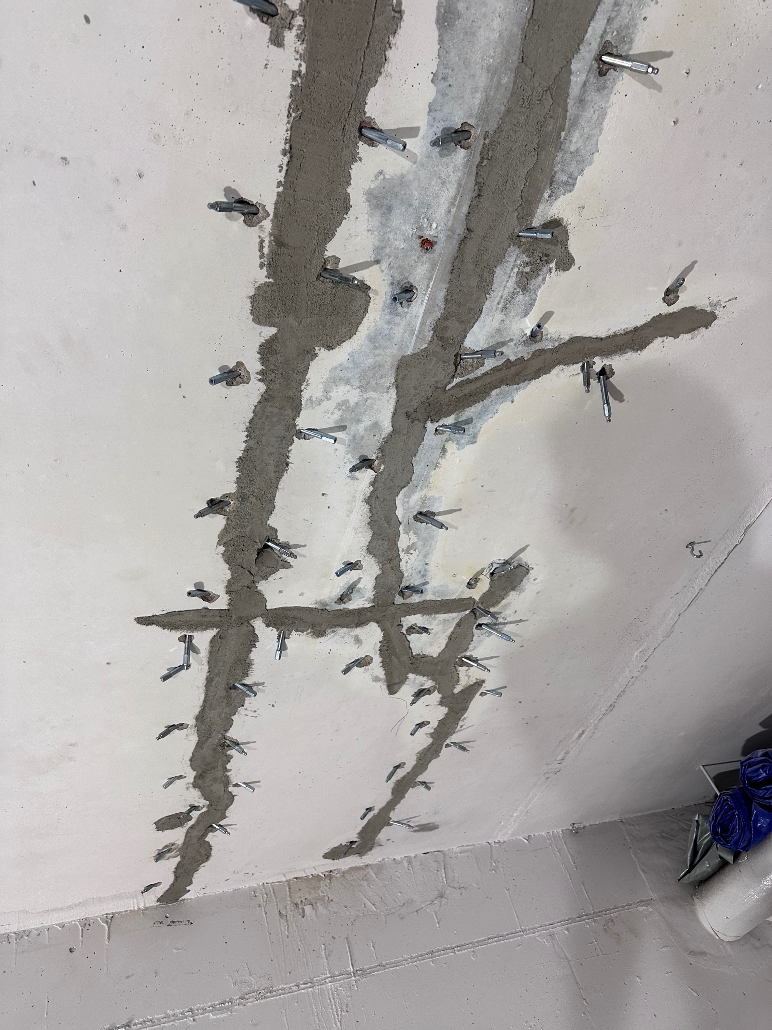 Betonowa ściana z widocznymi pęknięciami wypełnionymi szarym materiałem uszczelniającym i metalowymi kotwami, ukazująca etap naprawy konstrukcji.