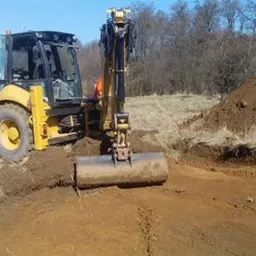 Żółto-czarna koparko-ładowarka w trakcie prac ziemnych, widoczny wykop w ziemi, usypana pryzma ziemi, drzewa w tle, słoneczny dzień.
