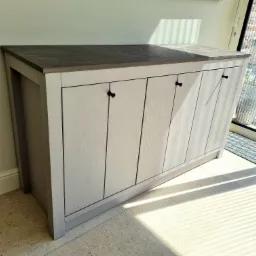 Biała szafka z drewnianymi frontami i ciemnym blatem, z czarnymi uchwytami, stojąca na jasnej podłodze w pobliżu okna z widokiem na zewnątrz, oświetlona promieniami słońca.