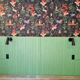 EWDAM Damian Kulpa - Zielona, ryflowana ściana z czarnymi lampkami nocnymi i gniazdkami elektrycznymi, nad nią tapeta w fantazyjne zwierzęta na ciemnym tle, całość dopełnia drewniana podłoga.