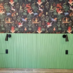 Zielona, ryflowana ściana z czarnymi lampkami nocnymi i gniazdkami elektrycznymi, nad nią tapeta w fantazyjne zwierzęta na ciemnym tle, całość dopełnia drewniana podłoga.