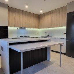 EWDAM Damian Kulpa - Nowoczesna kuchnia z wyspą i rozkładanym blatem, jasne fronty szafek, drewniane górne szafki, czarny sprzęt AGD, oświetlenie LED podszafkowe, widoczny fragment ściany z cegły.