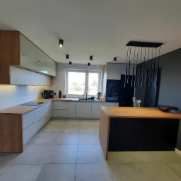 Nowoczesna kuchnia z szarymi szafkami i drewnianymi blatami, oświetlona punktowymi lampami sufitowymi i wiszącą lampą nad wyspą kuchenną, z elementami dekoracyjnymi z drewnianych listew na ścianie.