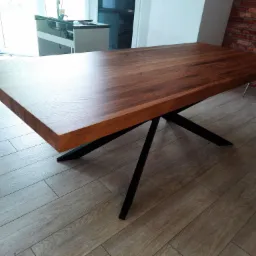 Prostokątny stół z blatem z ciemnego drewna i czarnymi, metalowymi nogami w kształcie gwiazdy, ustawiony na podłodze z paneli imitujących drewno. W tle fragment ściany z cegły i krzesło.