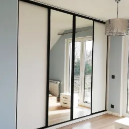 Nowoczesna szafa przesuwna z białymi frontami i lustrzanymi panelami w czarnych ramach, odbijająca fragment pokoju z oknem balkonowym i lampą wiszącą.