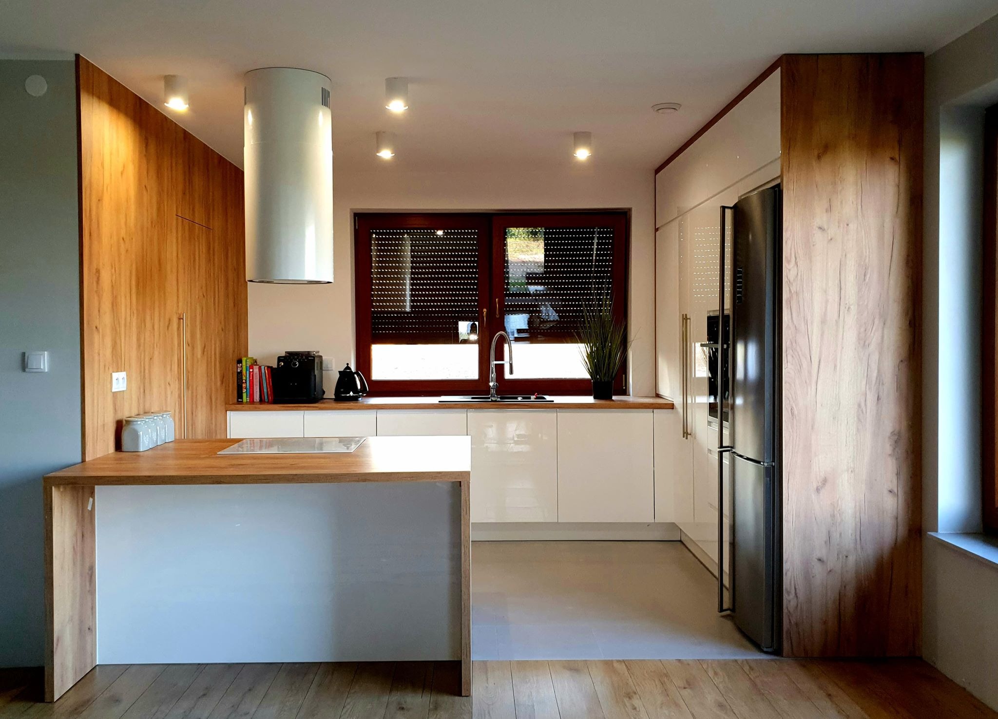 Nowoczesna kuchnia z białymi szafkami, drewnianymi akcentami, wyspą kuchenną i dużym oknem z żaluzjami.