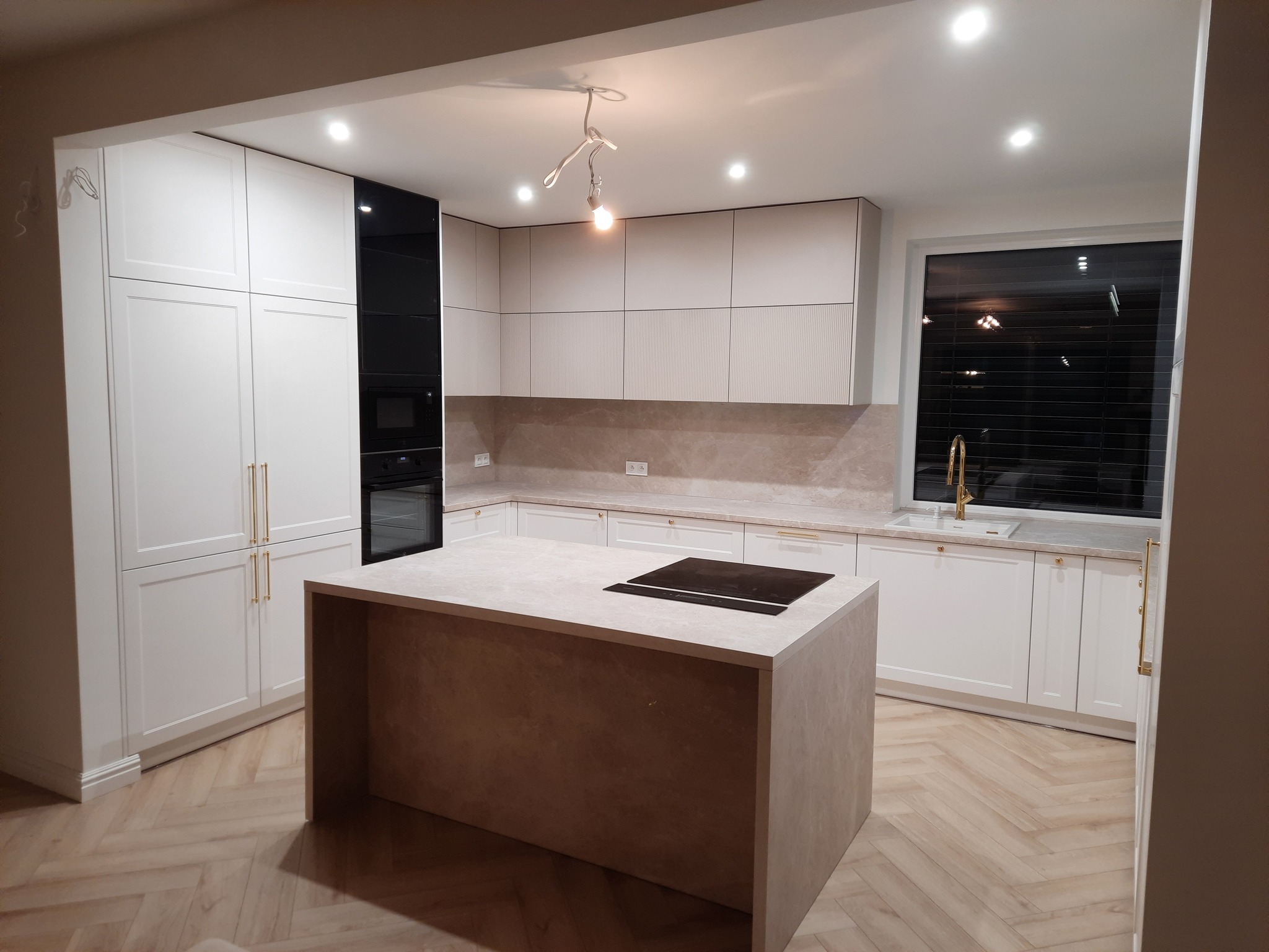 Nowoczesna kuchnia z wyspą, białe szafki z frezowanymi frontami i złotymi uchwytami, czarny panel z piekarnikiem, drewniana podłoga w jodełkę, oświetlenie punktowe i lampa wisząca z żarówką.