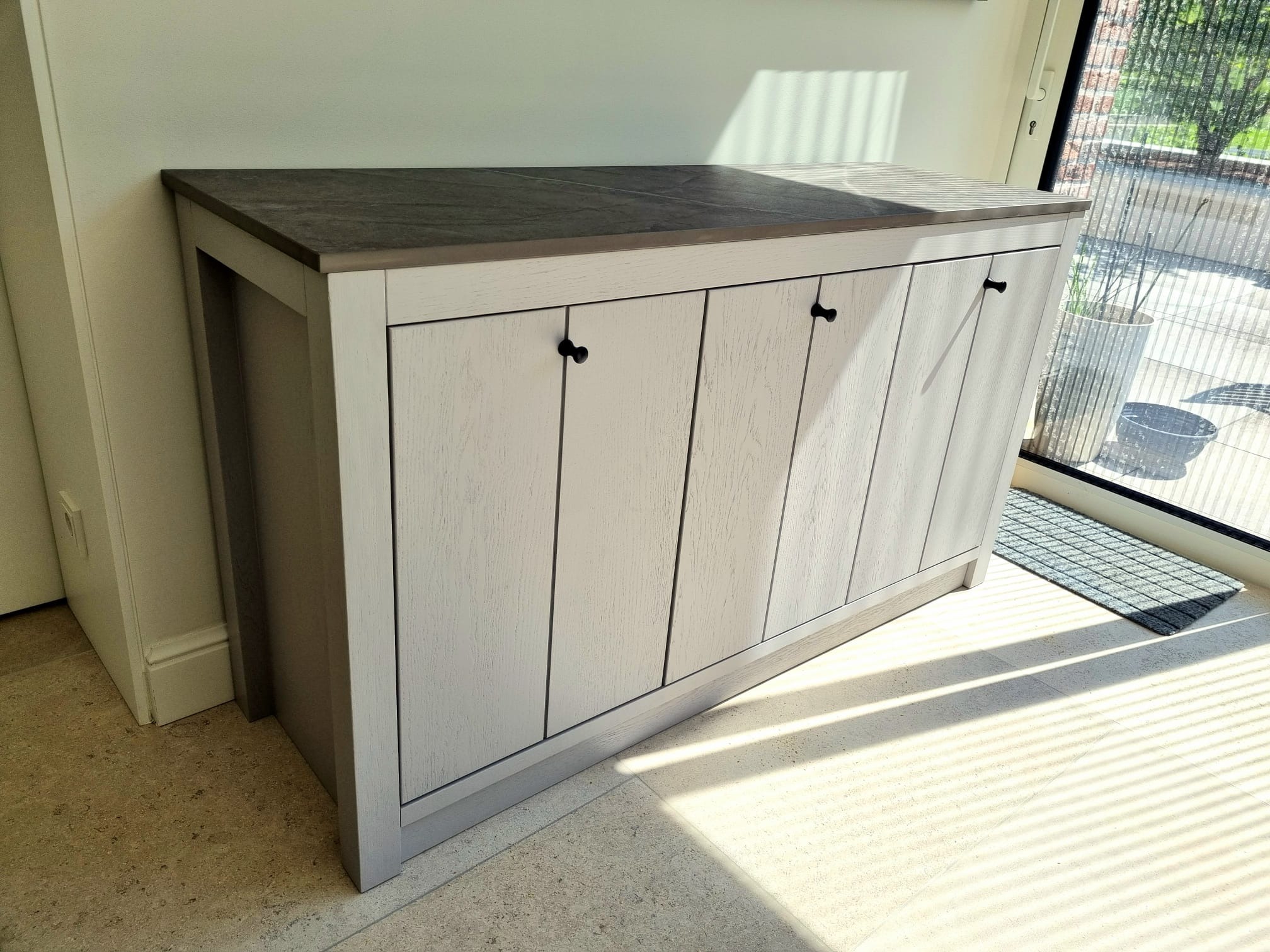 Biała szafka z drewnianymi frontami i ciemnym blatem, z czarnymi uchwytami, stojąca na jasnej podłodze w pobliżu okna z widokiem na zewnątrz, oświetlona promieniami słońca.