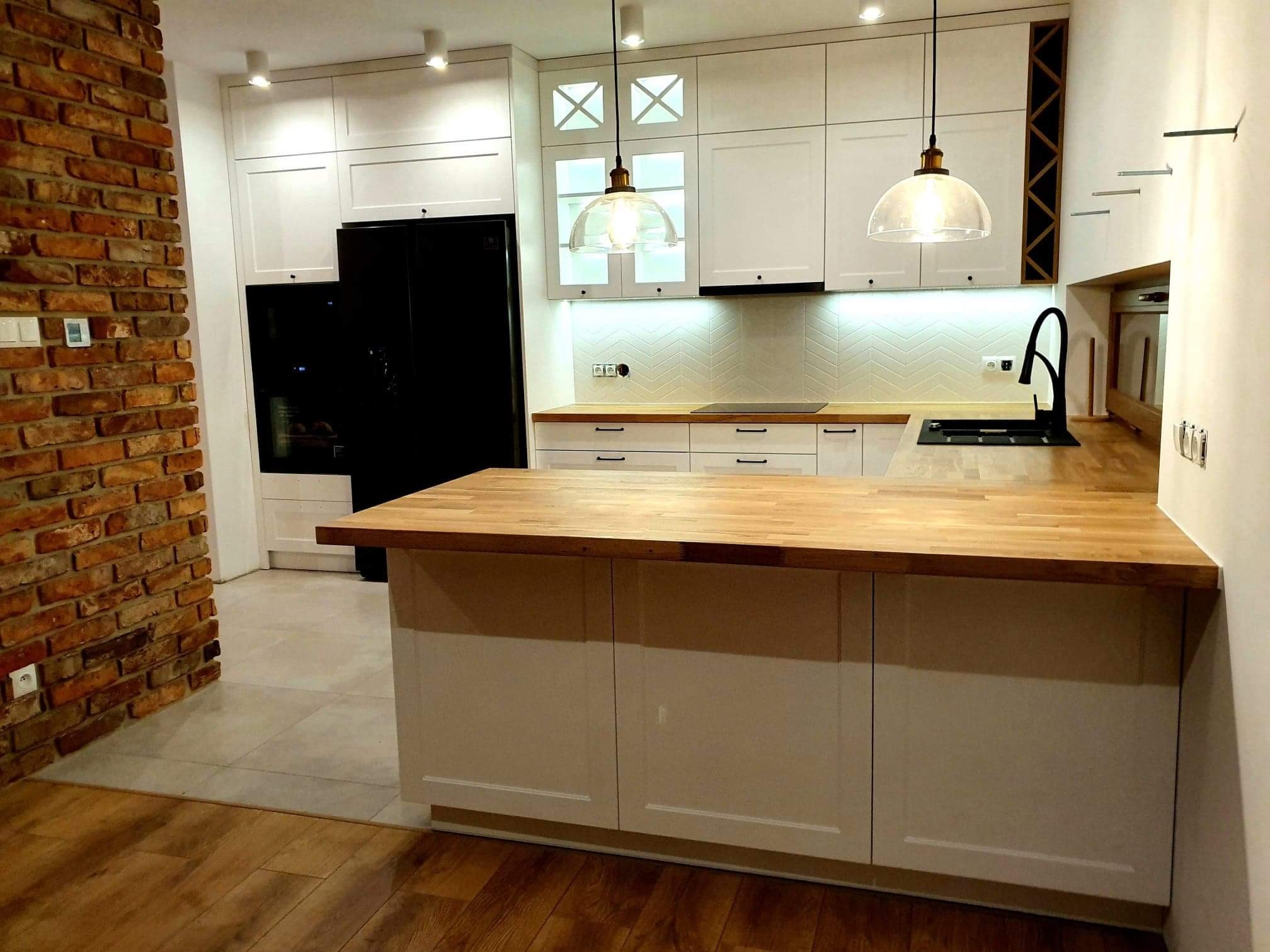 Nowoczesna kuchnia z białą zabudową, drewnianym blatem, ceglaną ścianą i wiszącymi lampami z kloszami