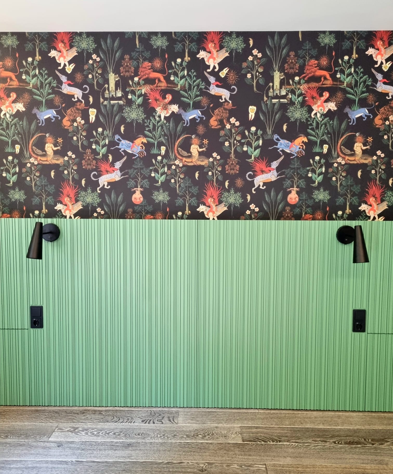 Zielona, ryflowana ściana z czarnymi lampkami nocnymi i gniazdkami elektrycznymi, nad nią tapeta w fantazyjne zwierzęta na ciemnym tle, całość dopełnia drewniana podłoga.