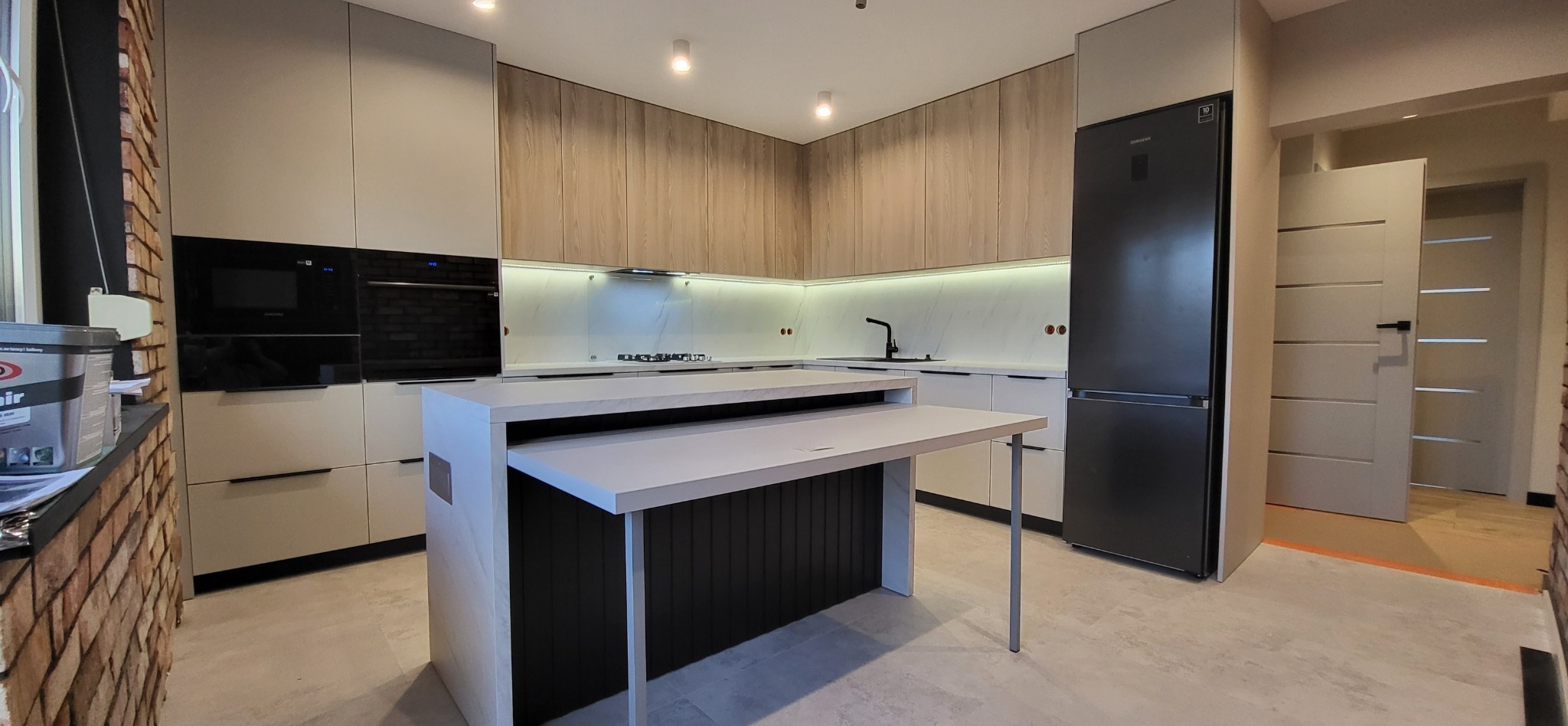 Nowoczesna kuchnia z wyspą i rozkładanym blatem, jasne fronty szafek, drewniane górne szafki, czarny sprzęt AGD, oświetlenie LED podszafkowe, widoczny fragment ściany z cegły.