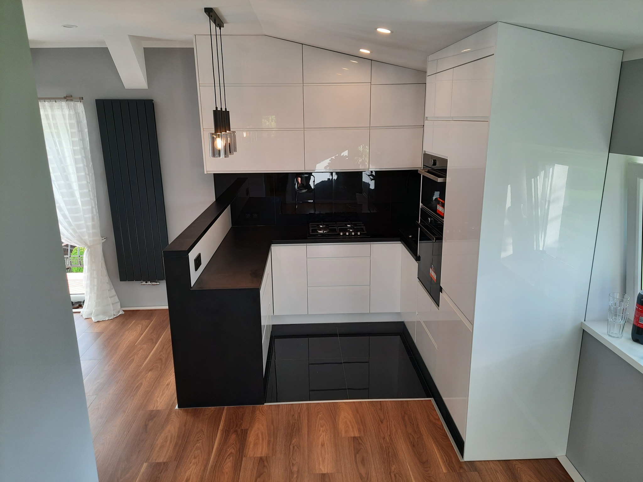 Nowoczesna kuchnia z białymi, lakierowanymi frontami szafek i czarnym blatem, oświetlona wiszącą lampą z trzema szklanymi kloszami, widok z perspektywy wejścia do pomieszczenia.