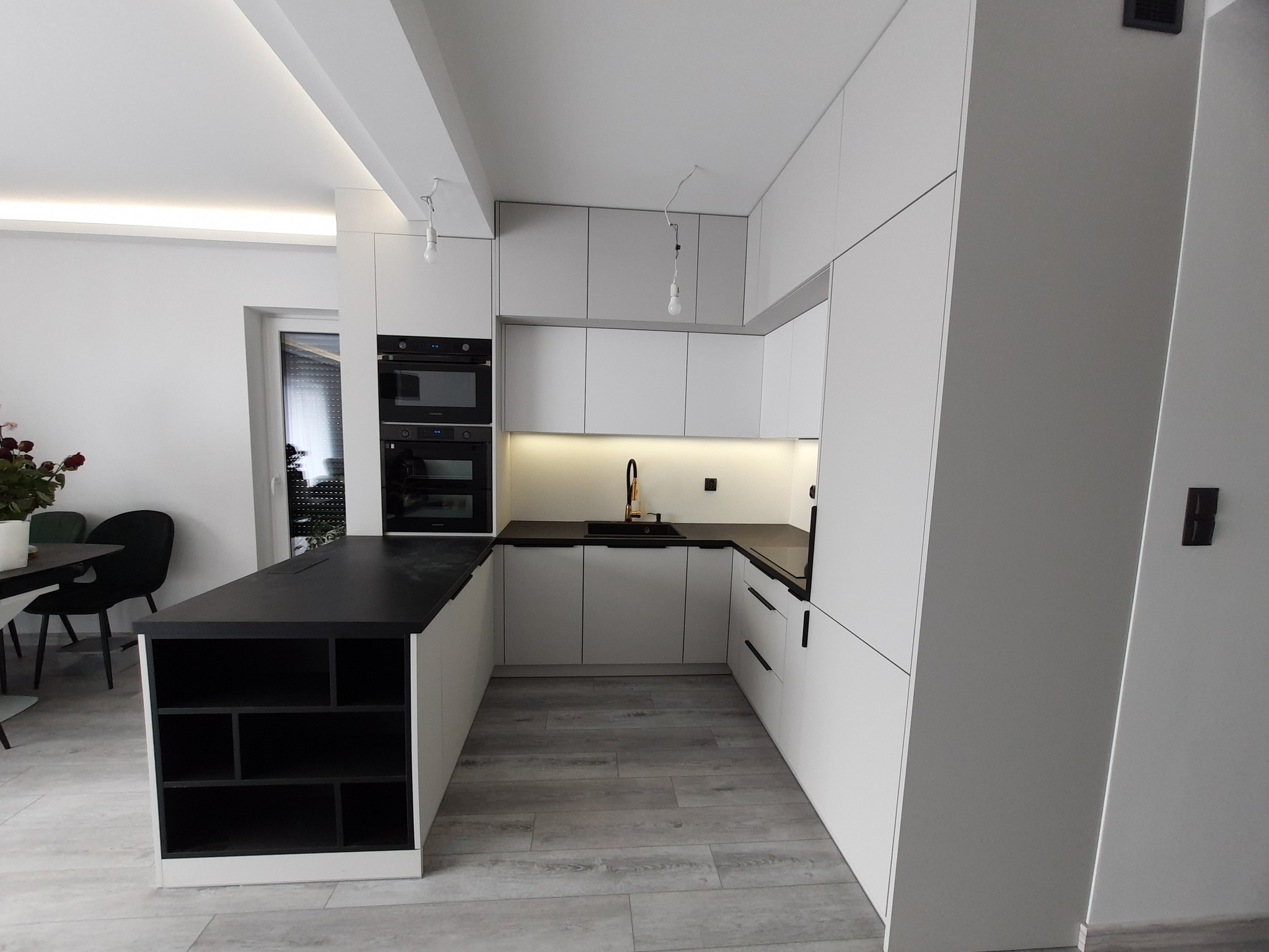 Nowoczesna kuchnia z białymi szafkami i czarnym blatem, oświetleniem LED pod szafkami wiszącymi oraz wyspą z półkami skierowanymi do salonu.