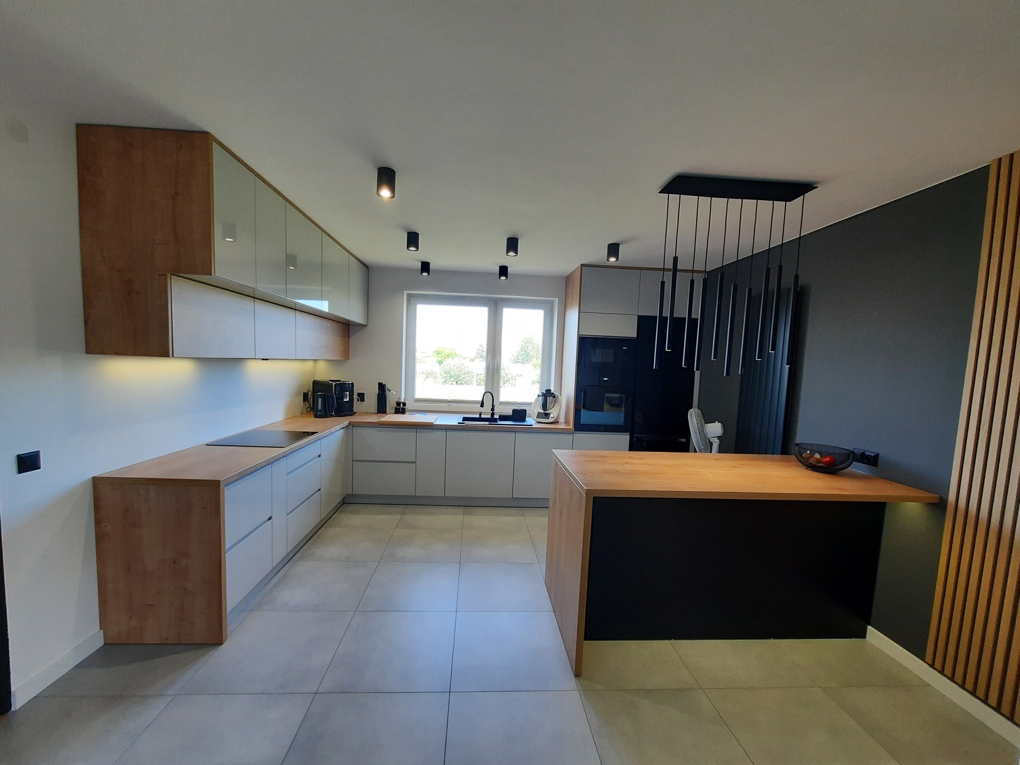 Nowoczesna kuchnia z szarymi szafkami i drewnianymi blatami, oświetlona punktowymi lampami sufitowymi i wiszącą lampą nad wyspą kuchenną, z elementami dekoracyjnymi z drewnianych listew na ścianie.