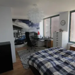 Sypialnia z fototapetą przedstawiającą stację benzynową i klasyczny samochód, biurko z krzesłem obrotowym, biała lampa wisząca w kształcie kuli i łóżko z niebiesko-białą pościelą w kratę.