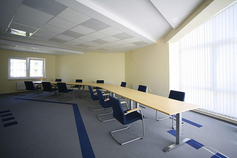 Sala konferencyjna z długim, jasnym stołem i niebieskimi krzesłami, szary dywan z niebieskimi akcentami, okno z żaluzjami pionowymi.