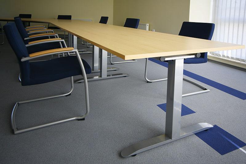 Długa, jasna sala konferencyjna z dużym stołem z jasnego drewna na metalowych nogach i ciemnoniebieskimi krzesłami z drewnianymi podłokietnikami, szara wykładzina z niebieskimi akcentami.