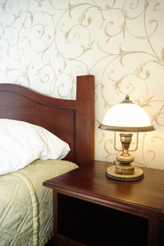 Fragment sypialni z drewnianym łóżkiem, wzorzystą tapetą i lampką nocną w stylu retro na ciemnobrązowej szafce nocnej.
