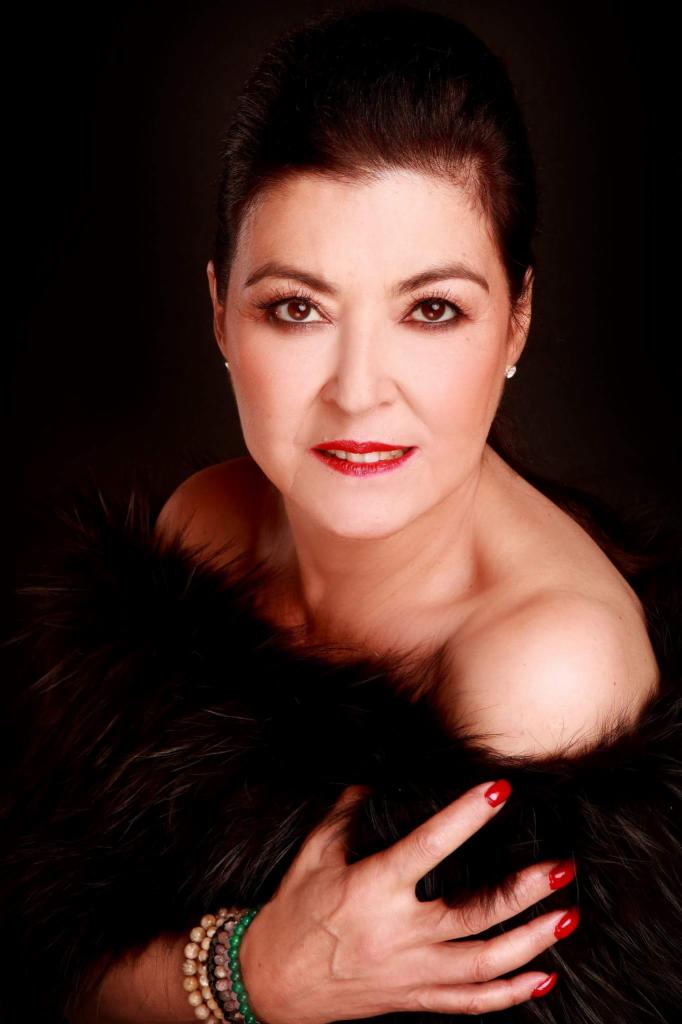 Portret kobiety z ciemnymi włosami, czerwonymi ustami i paznokciami, owiniętej w czarne futro, z bransoletką z koralików na nadgarstku, na czarnym tle.