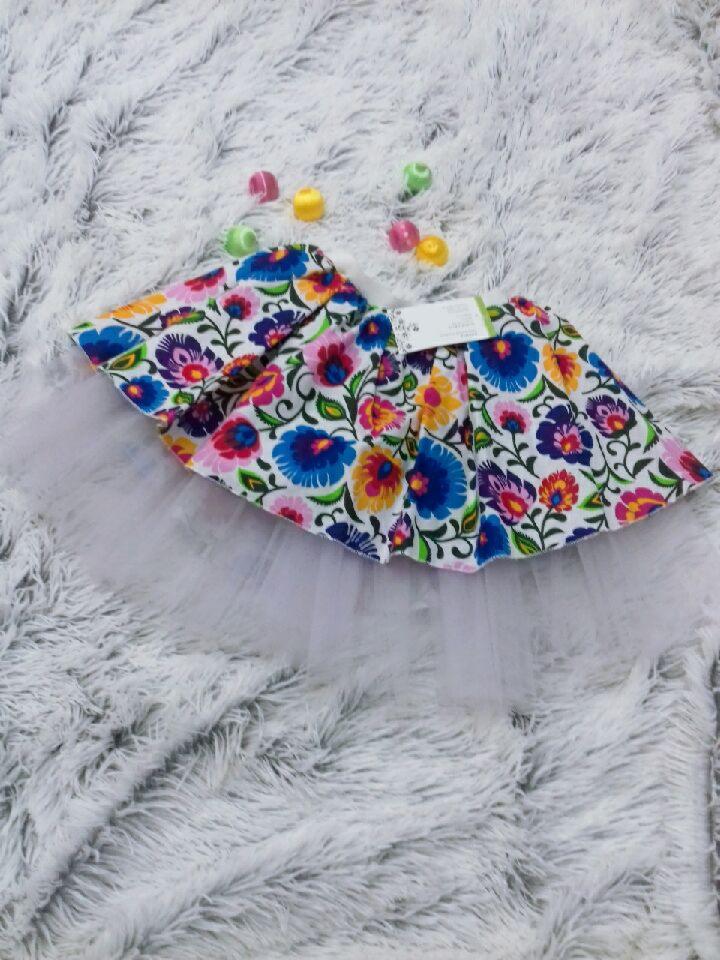 Dziecięca spódniczka tutu z folkowym wzorem kwiatowym, leżąca na puszystym, białym dywanie, ozdobiona kolorowymi, okrągłymi cukierkami.