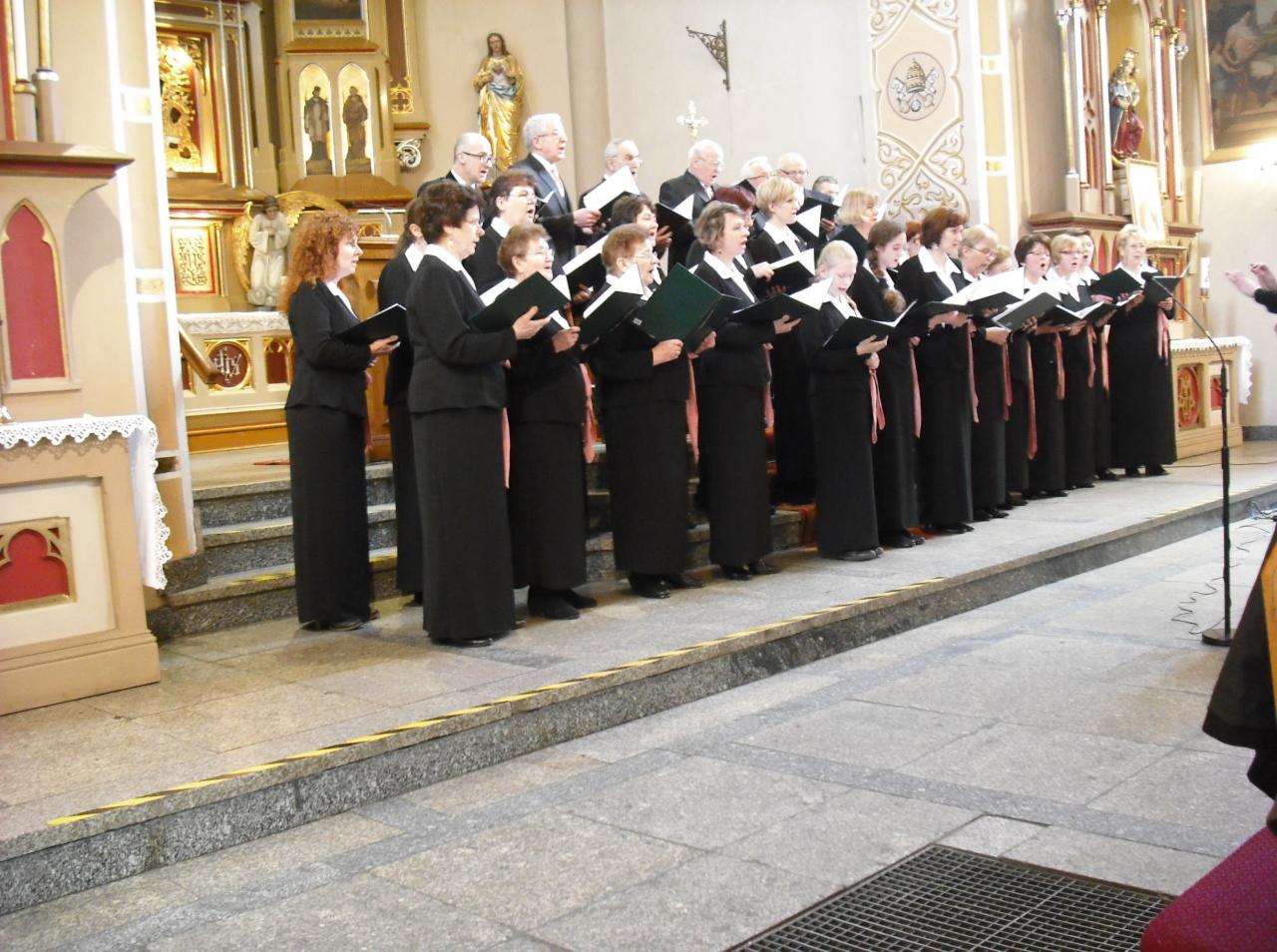 Chór kościelny w długich, czarnych szatach z różowymi szarfami, śpiewający z nut w kościele, z dyrygentem po prawej stronie.