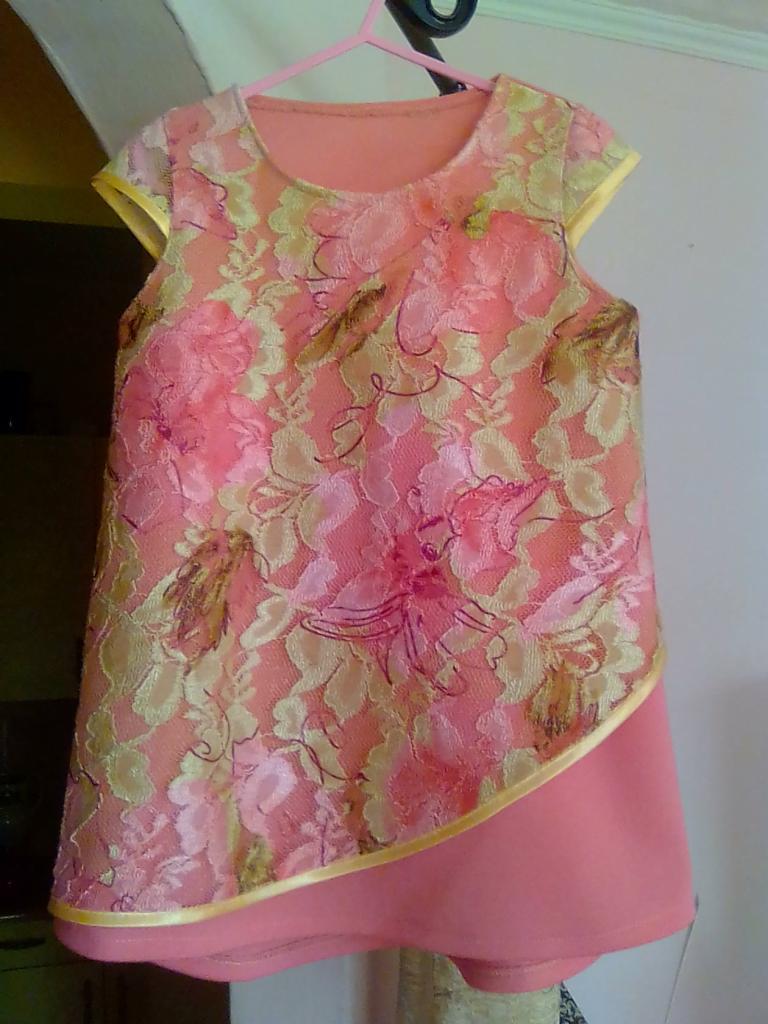 Różowa sukienka dziecięca z koronkową górą w kwiaty, wisząca na wieszaku z różowego tworzywa.