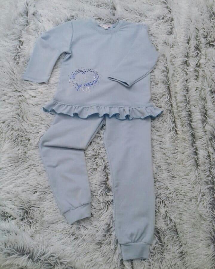 Jasnoniebieski komplet dziecięcy: bluza z długim rękawem i spodnie dresowe, ozdobione motywem serca z cekinów i falbanką, leżący na puszystym, jasnym tle.