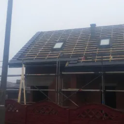 Dom w trakcie prac dekarskich: montaż łat i częściowe pokrycie dachówką w pochmurny dzień, widoczne rusztowania i okna dachowe.