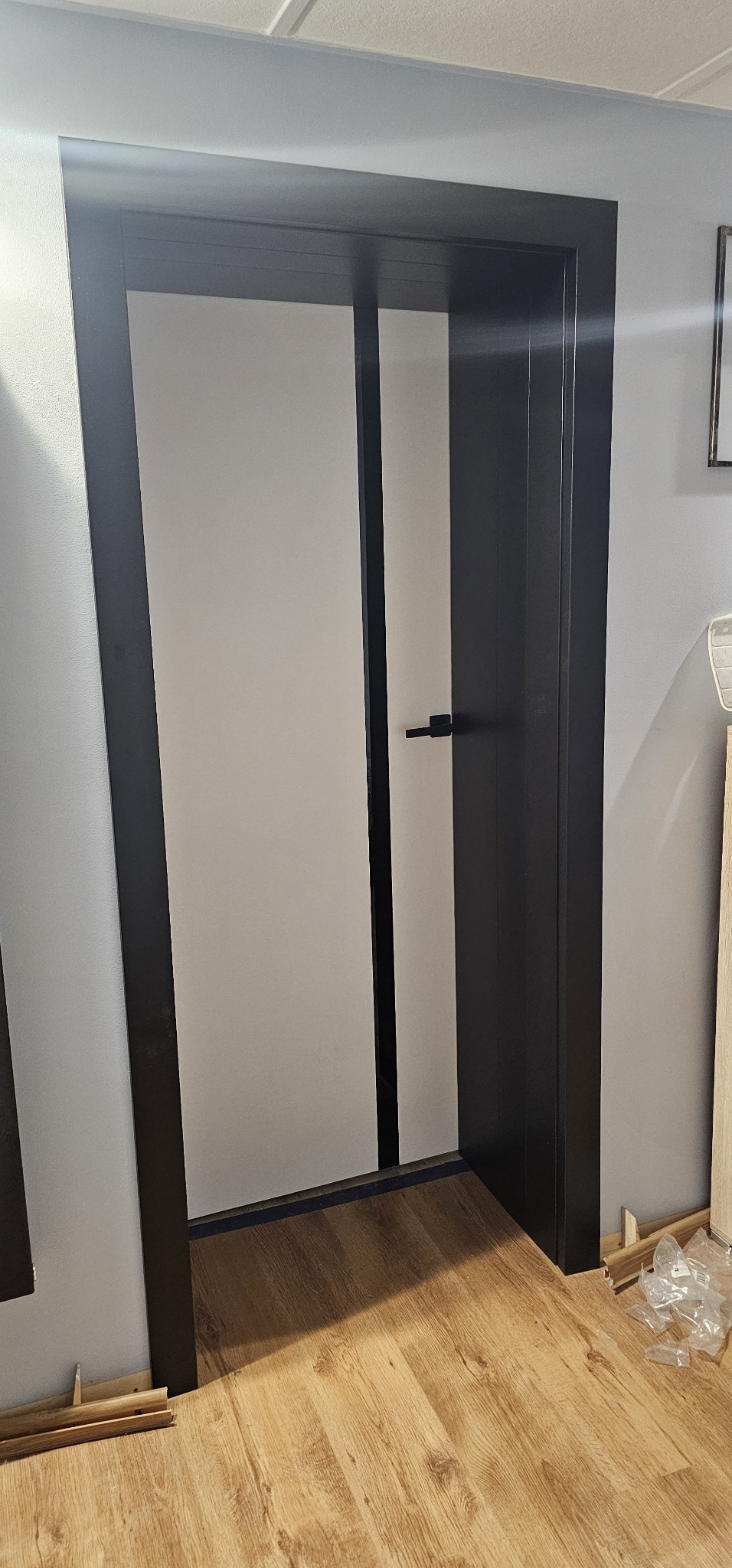 Zamknięte, białe drzwi z czarną ościeżnicą i klamką, widoczne na tle szarej ściany i drewnopodobnej podłogi z elementami montażowymi.