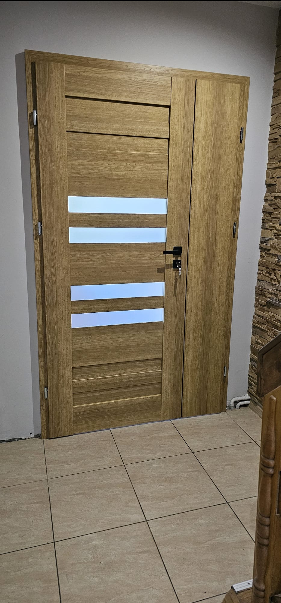 Zamknięte, dwuskrzydłowe drzwi wewnętrzne z drewnopodobną okleiną i trzema poziomymi przeszkleniami, widoczne zamek z kluczem, na podłodze jasne płytki, w tle ściana z dekoracyjnym kamieniem.