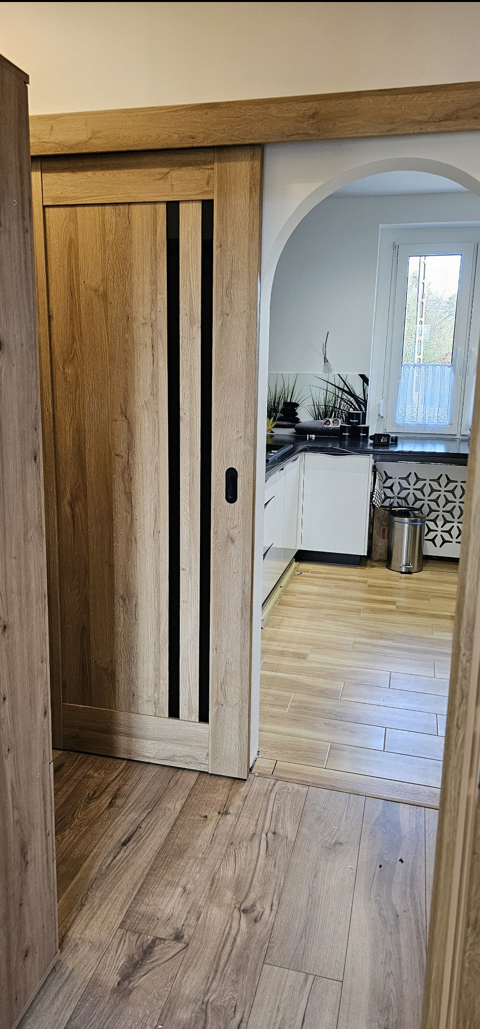 Drewniane drzwi przesuwne z czarnymi wstawkami, widok na kuchnię z białymi szafkami i oknem, podłoga wyłożona płytkami imitującymi drewno.