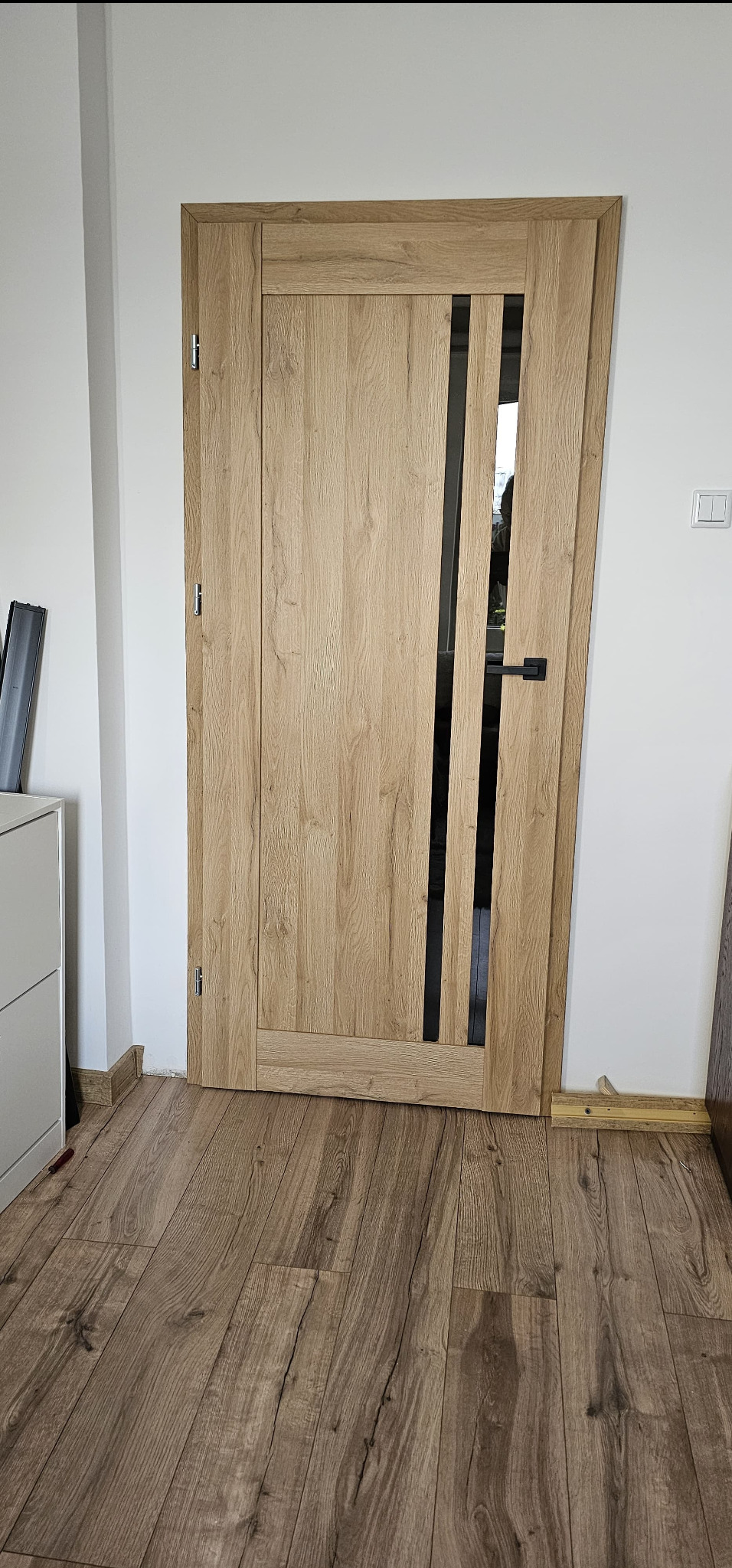 Jasne, drewnopodobne drzwi wewnętrzne z czarnymi, pionowymi wstawkami, zamontowane w białej ścianie, widok z perspektywy podłogi z paneli.