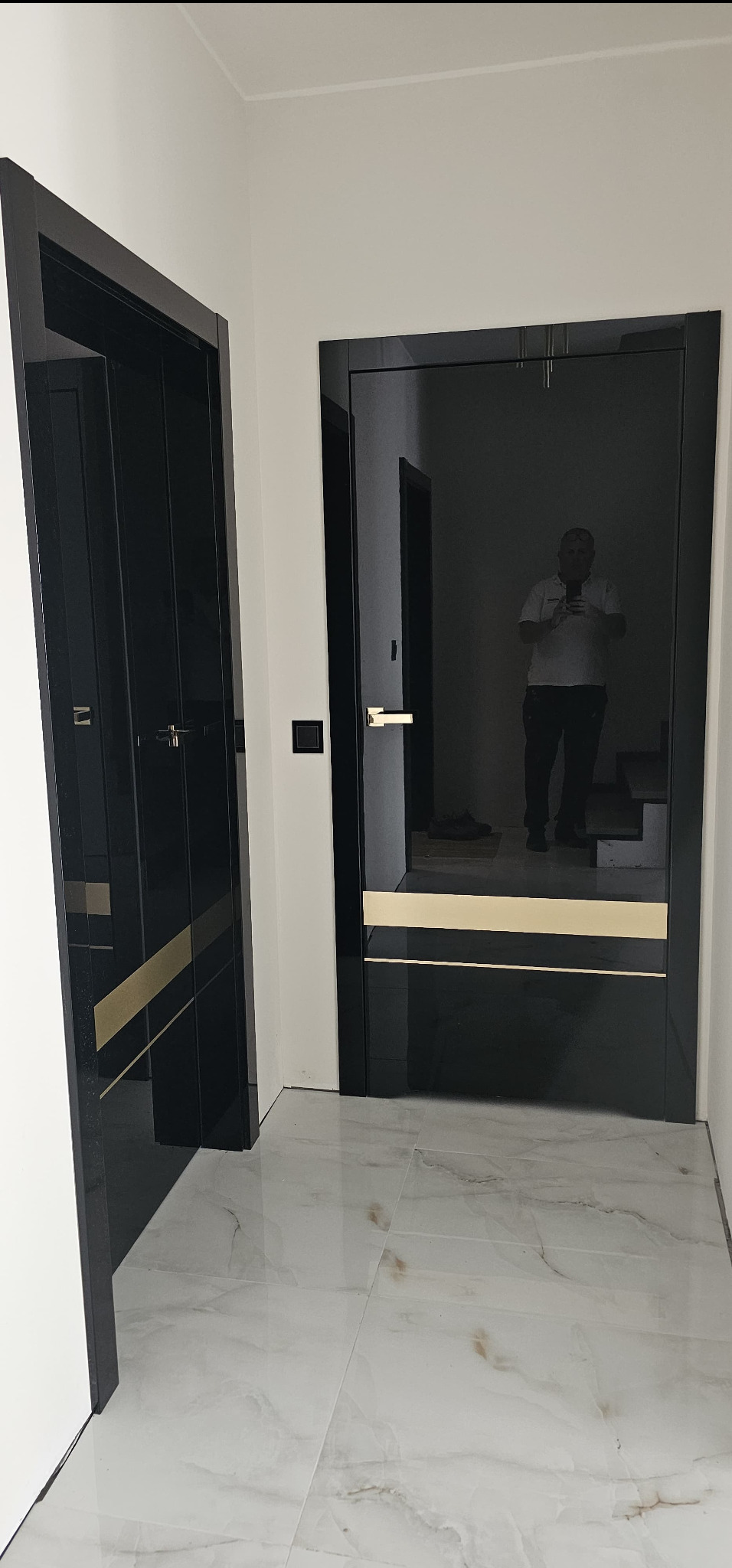 Dwa czarne drzwi wewnętrzne z złotymi akcentami, widoczne na tle białych ścian i marmurowej podłogi, z odbiciem osoby robiącej zdjęcie w powierzchni jednego z drzwi.