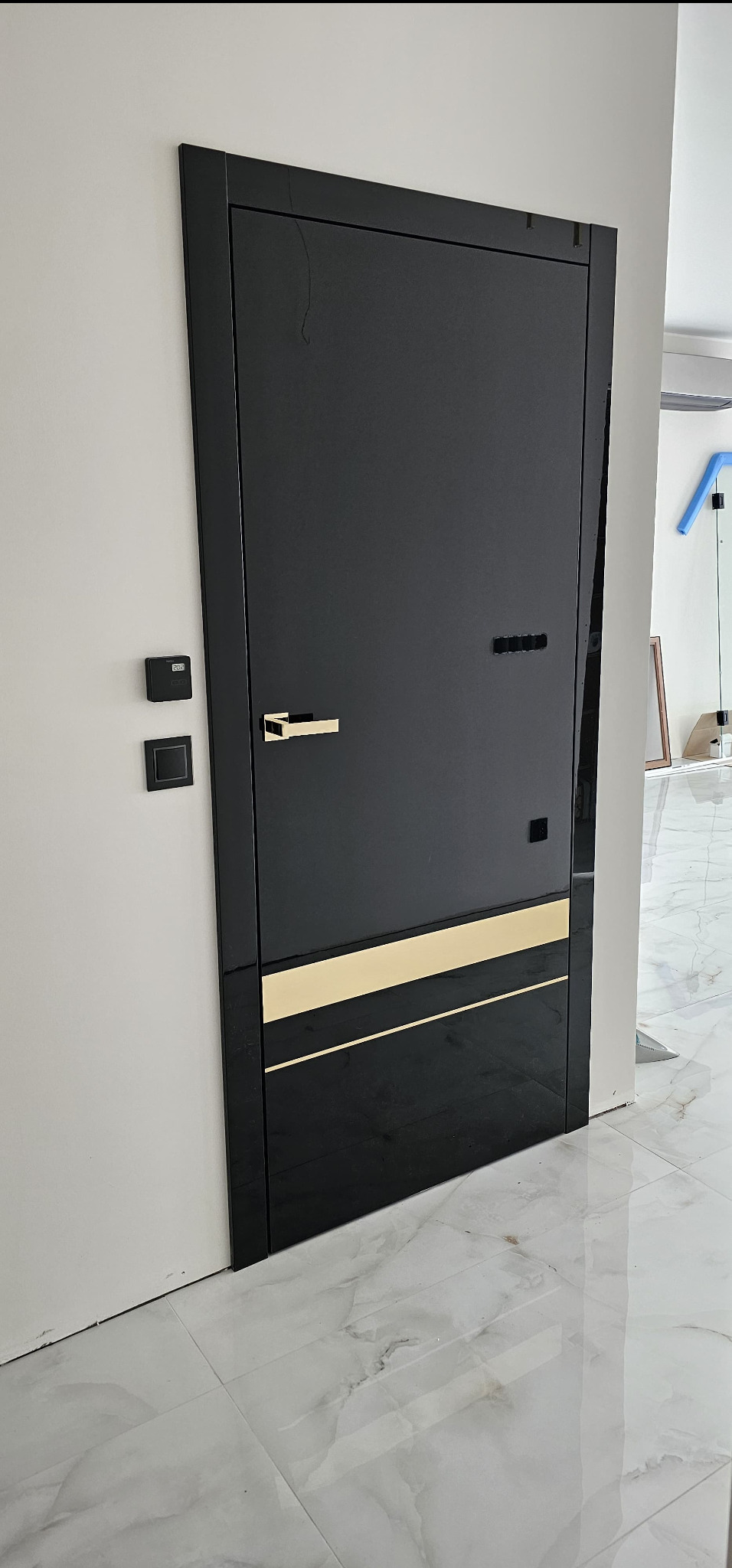 Czarne drzwi wewnętrzne z poziomymi złotymi paskami, zamontowane w nowoczesnym wnętrzu z marmurową podłogą i białymi ścianami, widoczny złoty uchwyt i czarny panel sterowania na ścianie.