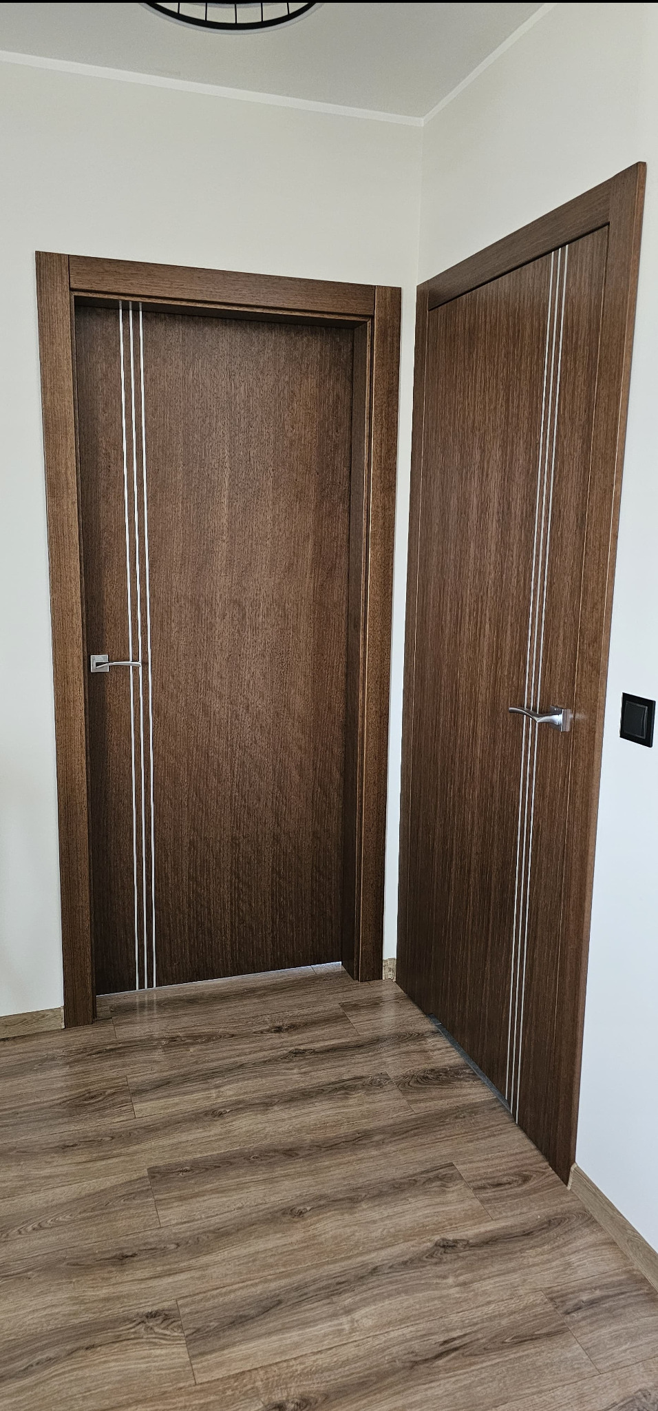 Dwa skrzydła drzwiowe w kolorze ciemnego drewna z trzema srebrnymi paskami, zamontowane w jasnym wnętrzu z drewnianą podłogą.