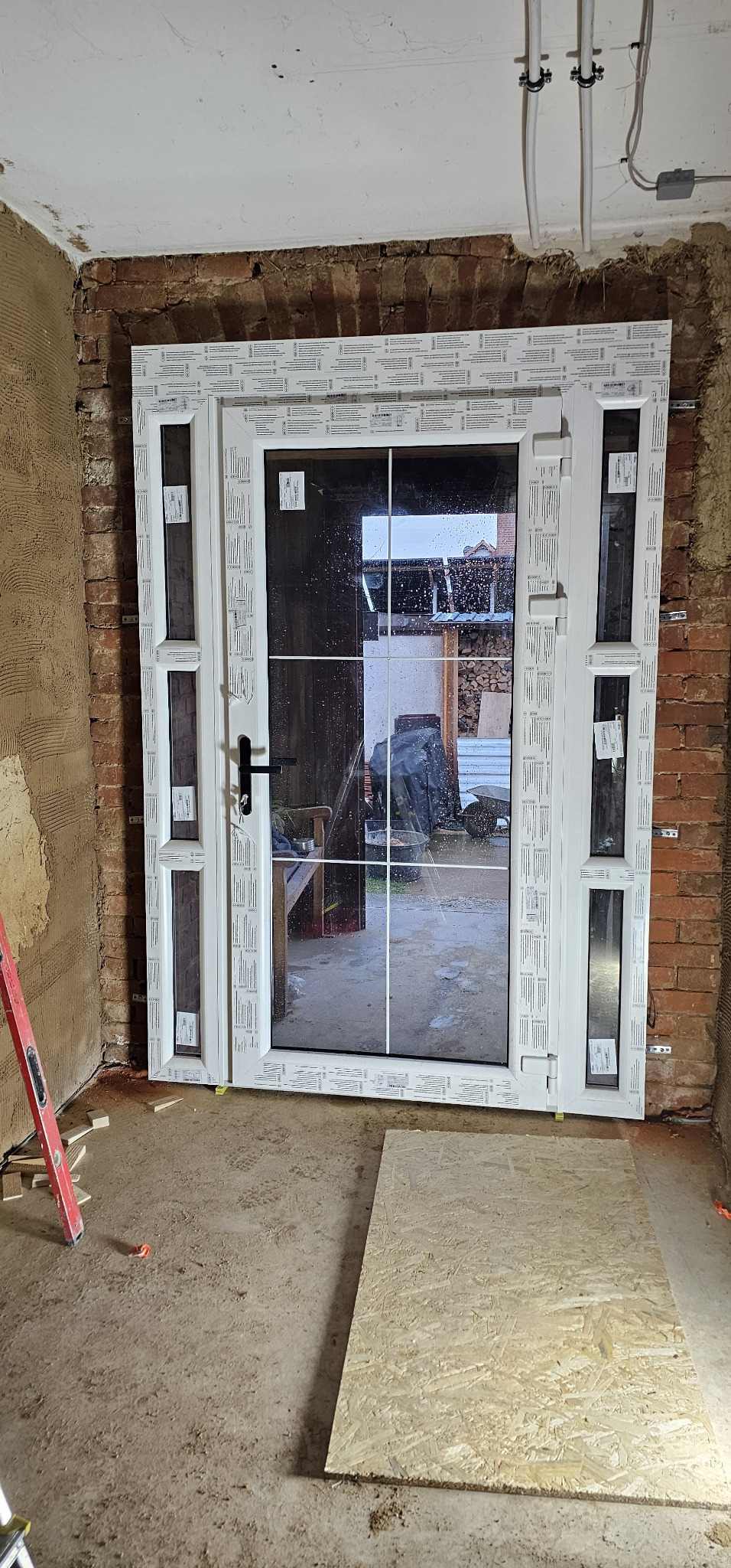 Białe, częściowo zamontowane drzwi z szybą i kratką, widoczne na tle surowej cegły i betonu. Elementy montażowe widoczne na ramie. Na zewnątrz widoczny zarys podwórka.