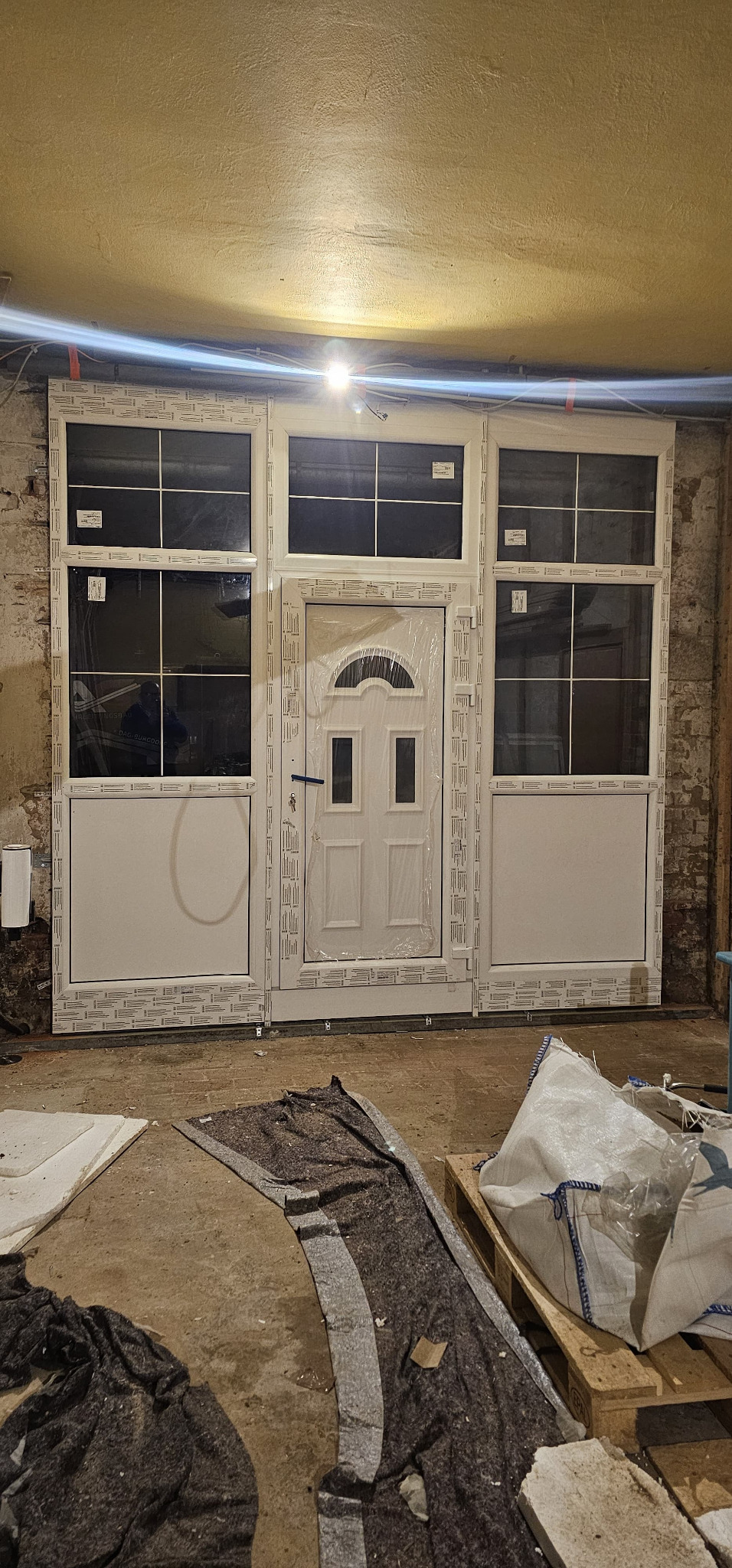 Białe drzwi wejściowe PCV z oknami bocznymi, jeszcze w folii ochronnej, zamontowane w surowym wnętrzu. Widoczne ślady prac montażowych.